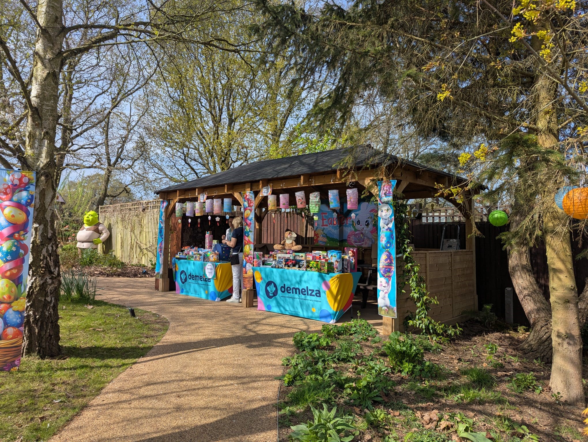 Wooden gazebo with Easter banners and lanterns, two tables with blue 'demelza' cloths displaying items, set in a garden with trees and a path.