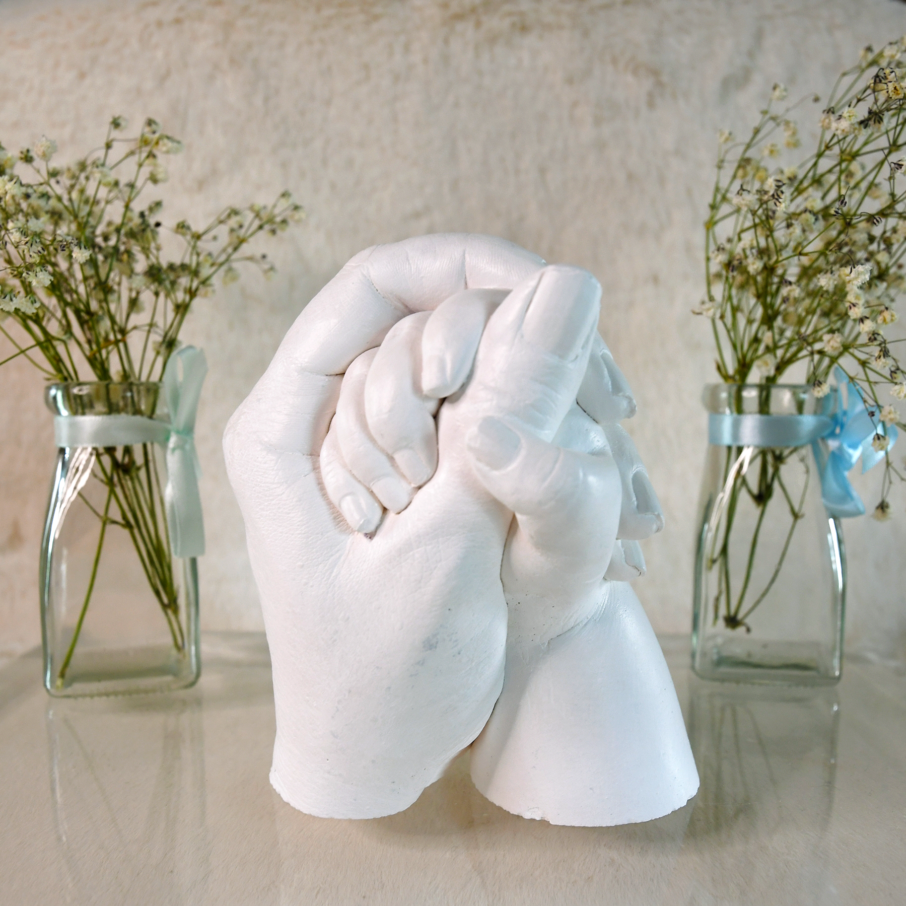 White plaster sculpture of two hands holding each other, flanked by glass vases with small white flowers tied with light blue ribbons.