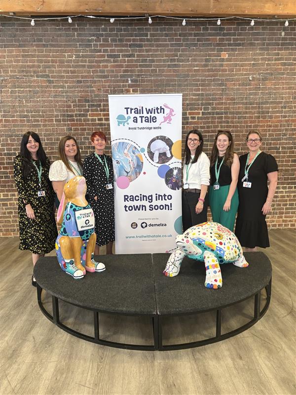 Six people stand by a "Trail with a Tale" banner and display featuring two colorful animal sculptures on a circular platform with "Racing into town soon!" text.
