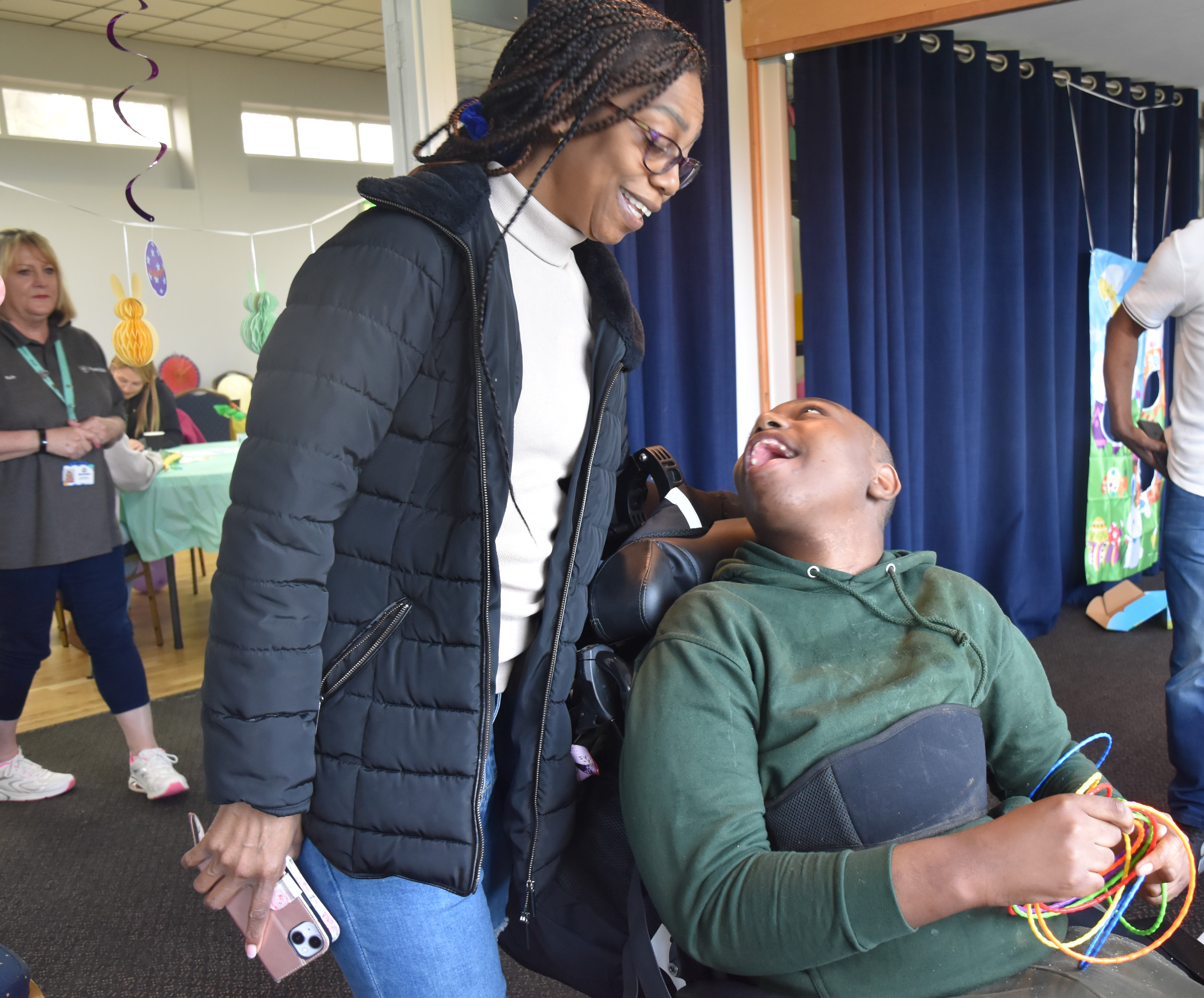 A woman with black hair looking down at a young boy in a wheel chair, both smiling at each other