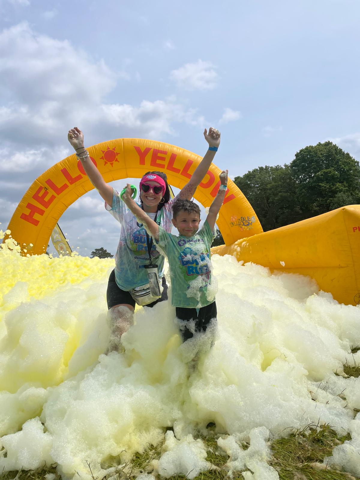 Adult and child run through yellow foam with arms raised, under a yellow inflatable arch reading 'HELLO YELLOW' during a Demelza Bubble Rush event.