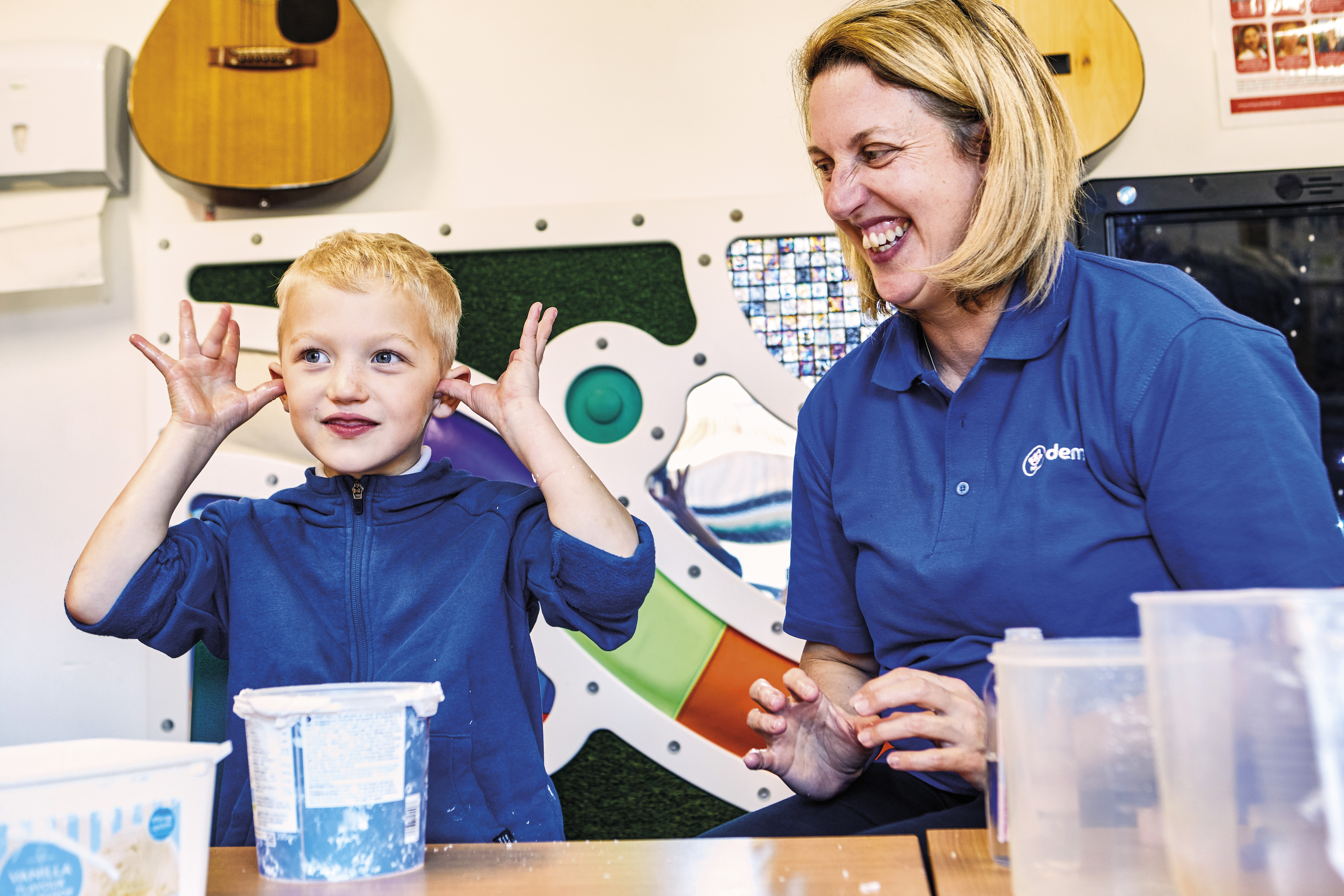A woman with blonde hair in a blue Demelza t-shirt smiling at a young boy with blonde hair with his fingers in his ears, smiling
