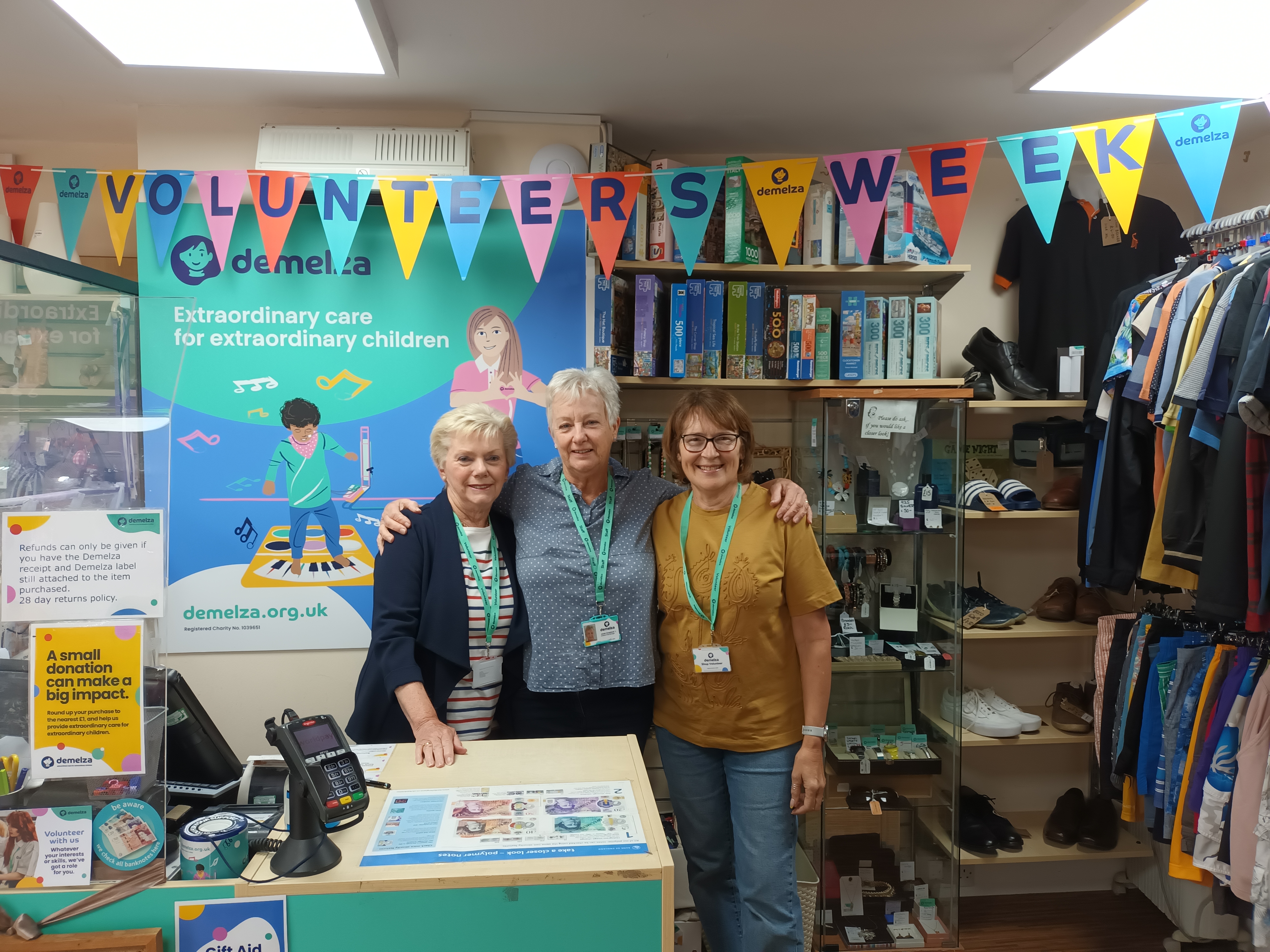 Three female volunteers stand behind the till in the Hythe shop, smiling amid colourful Volunteer’s Week 2025 banners.