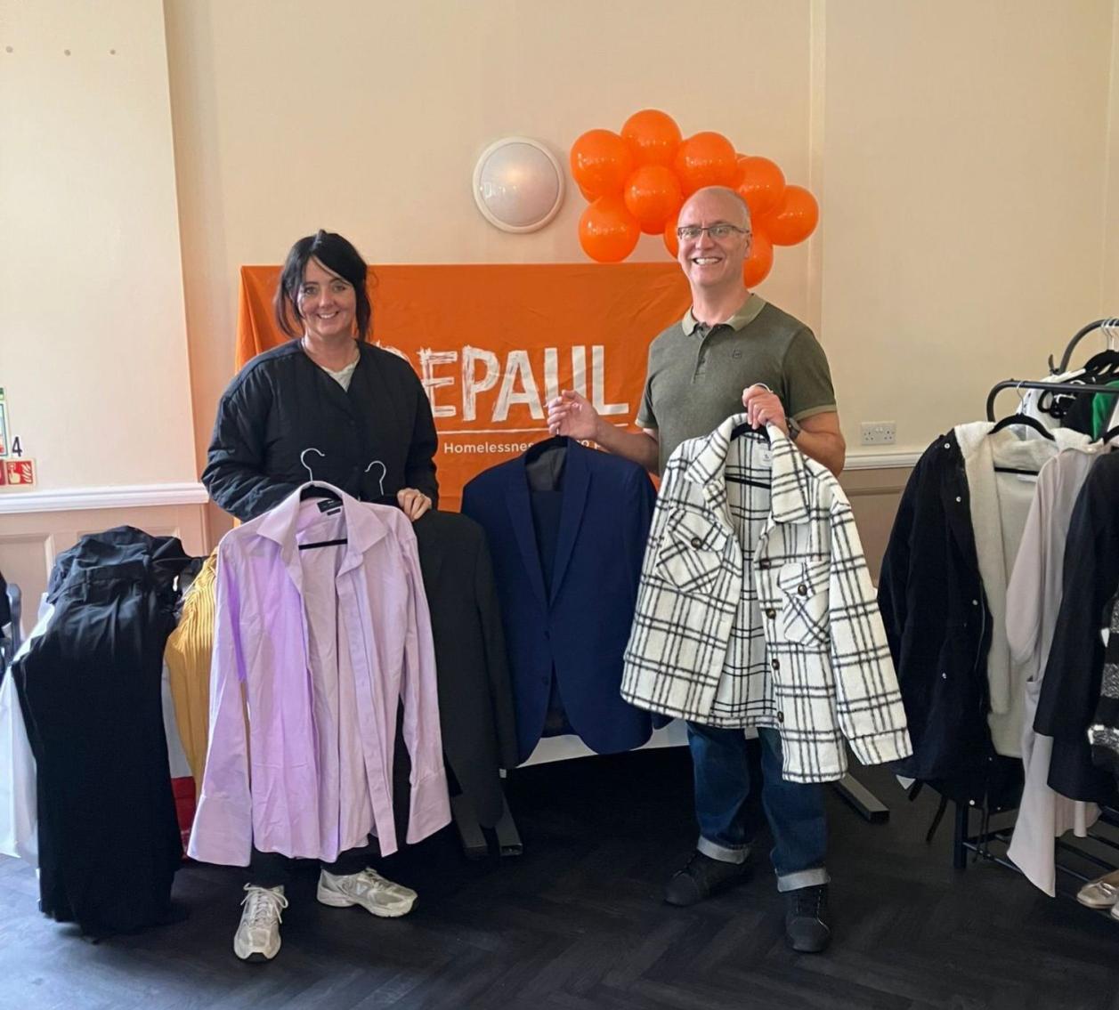 Two people hold clothes on hangers in front of an orange 'DEPAUL Homelessness' banner, with clothing racks on each side and orange balloons above.