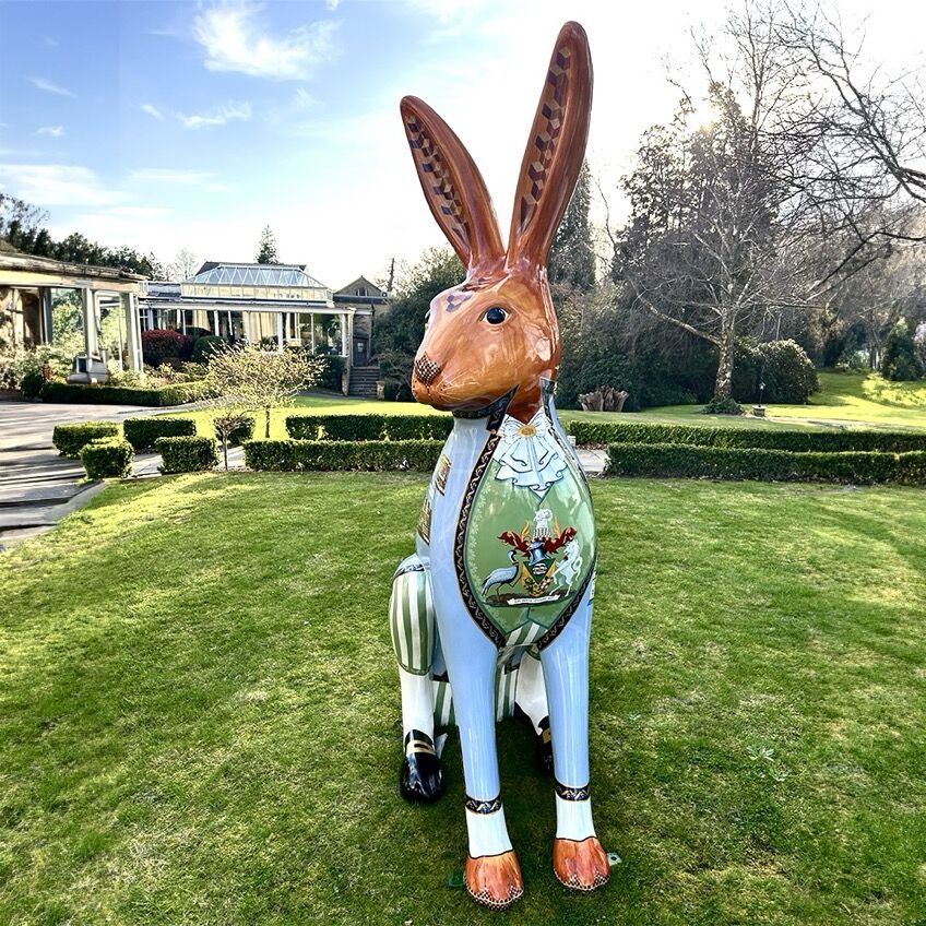 Large, colourful rabbit sculpture with intricate designs on a lawn, set against a garden with trimmed hedges and a building in the background.