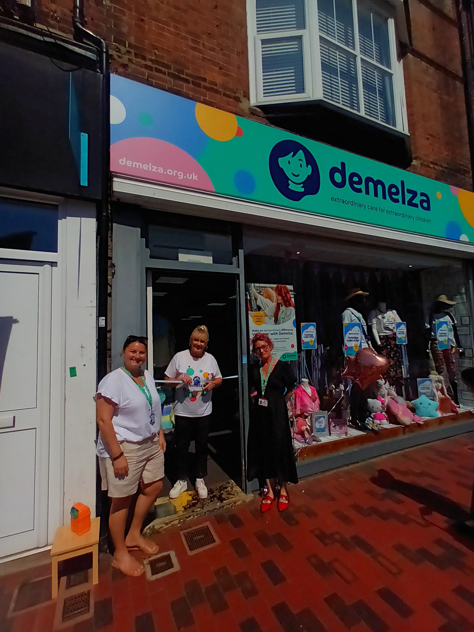 Three people outside a Demelza charity shop with a colorful sign and window display.