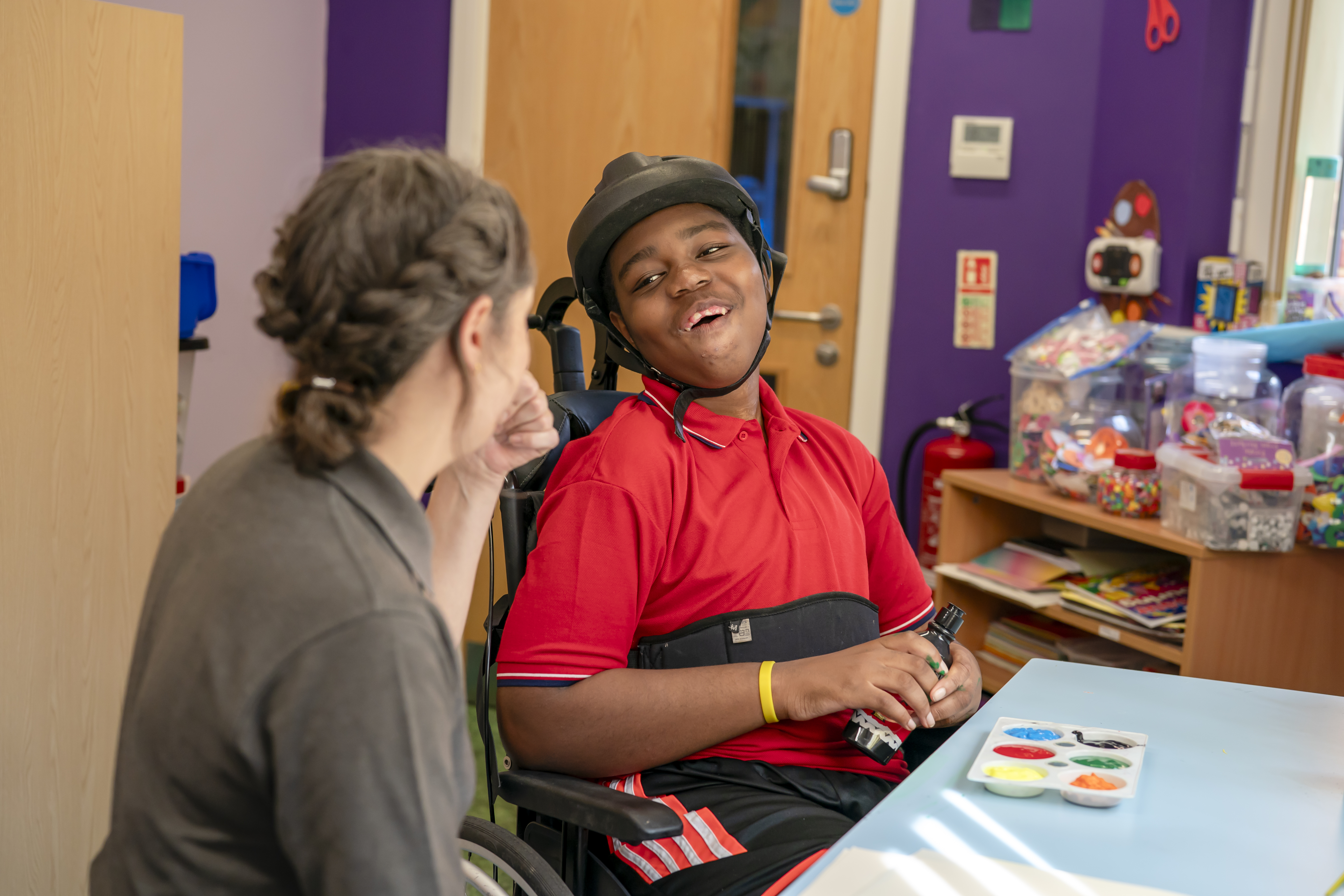A young boy with a helmet looking at a Demelza healthcare assistant in the art room