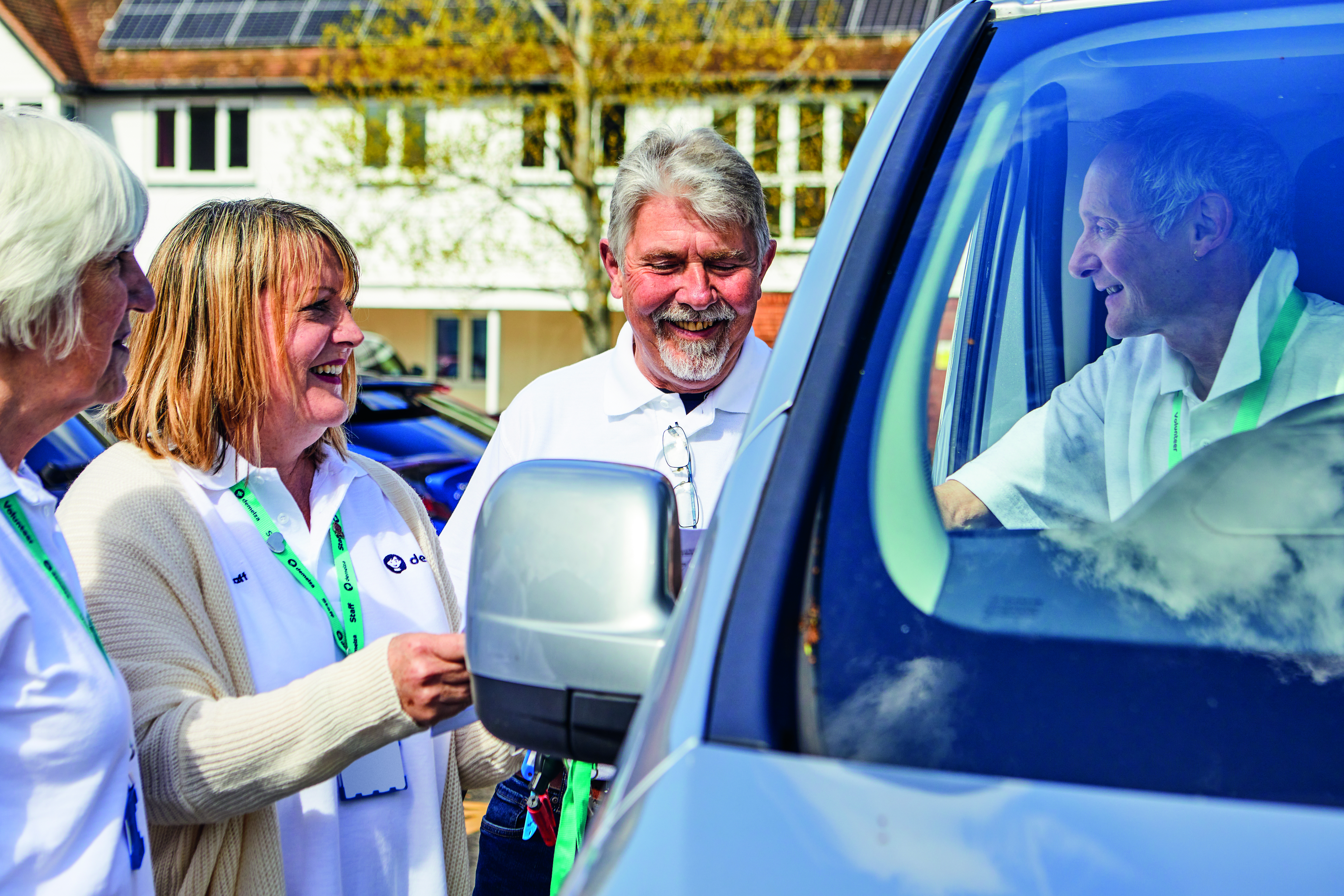 Four people wearing Demelza lanyards interacting near a Demelza vehicle; three stand outside while one is seated inside.