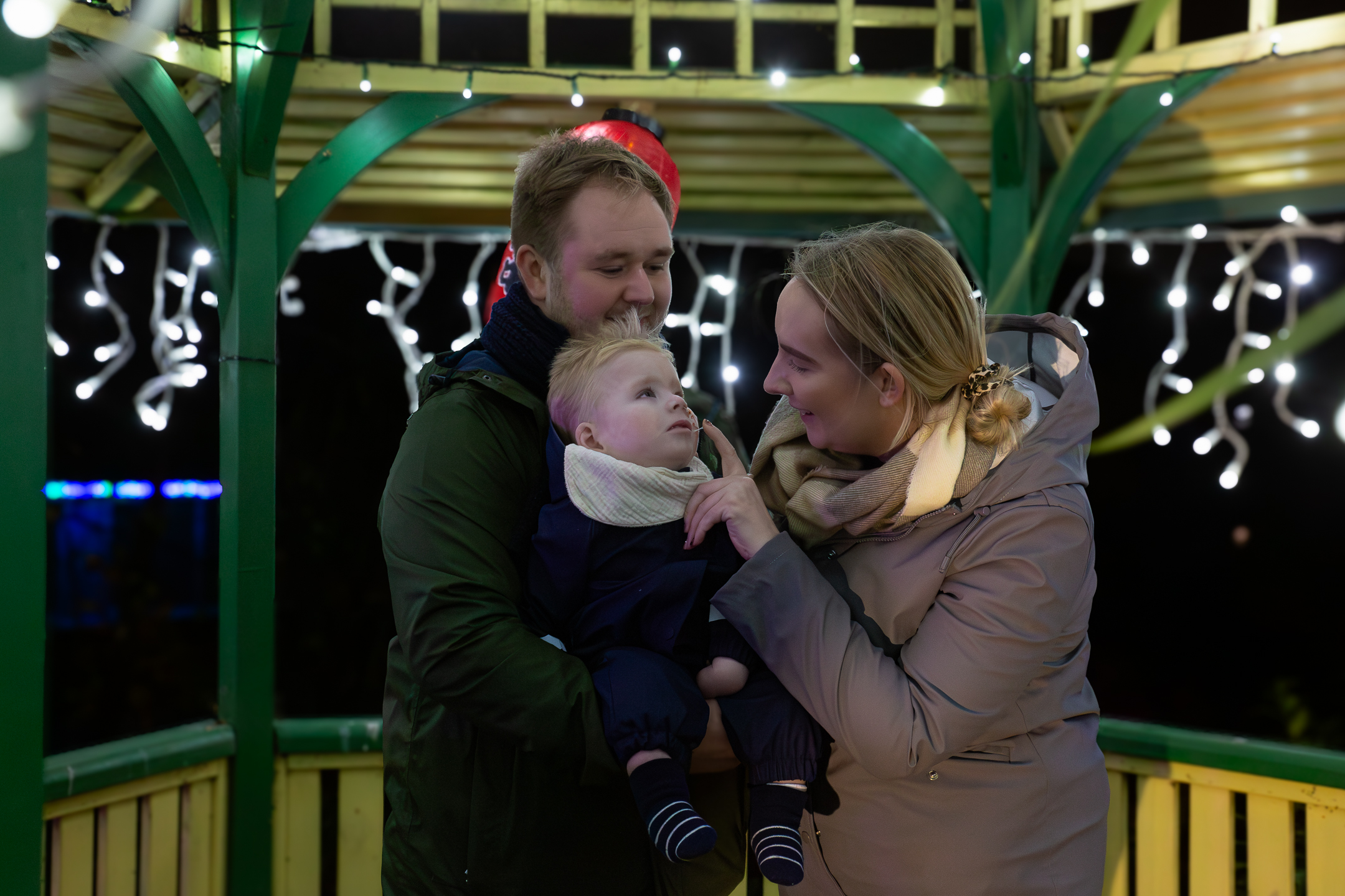 Fatehr and Mother stood under a gazebo with fairy lights holding their baby