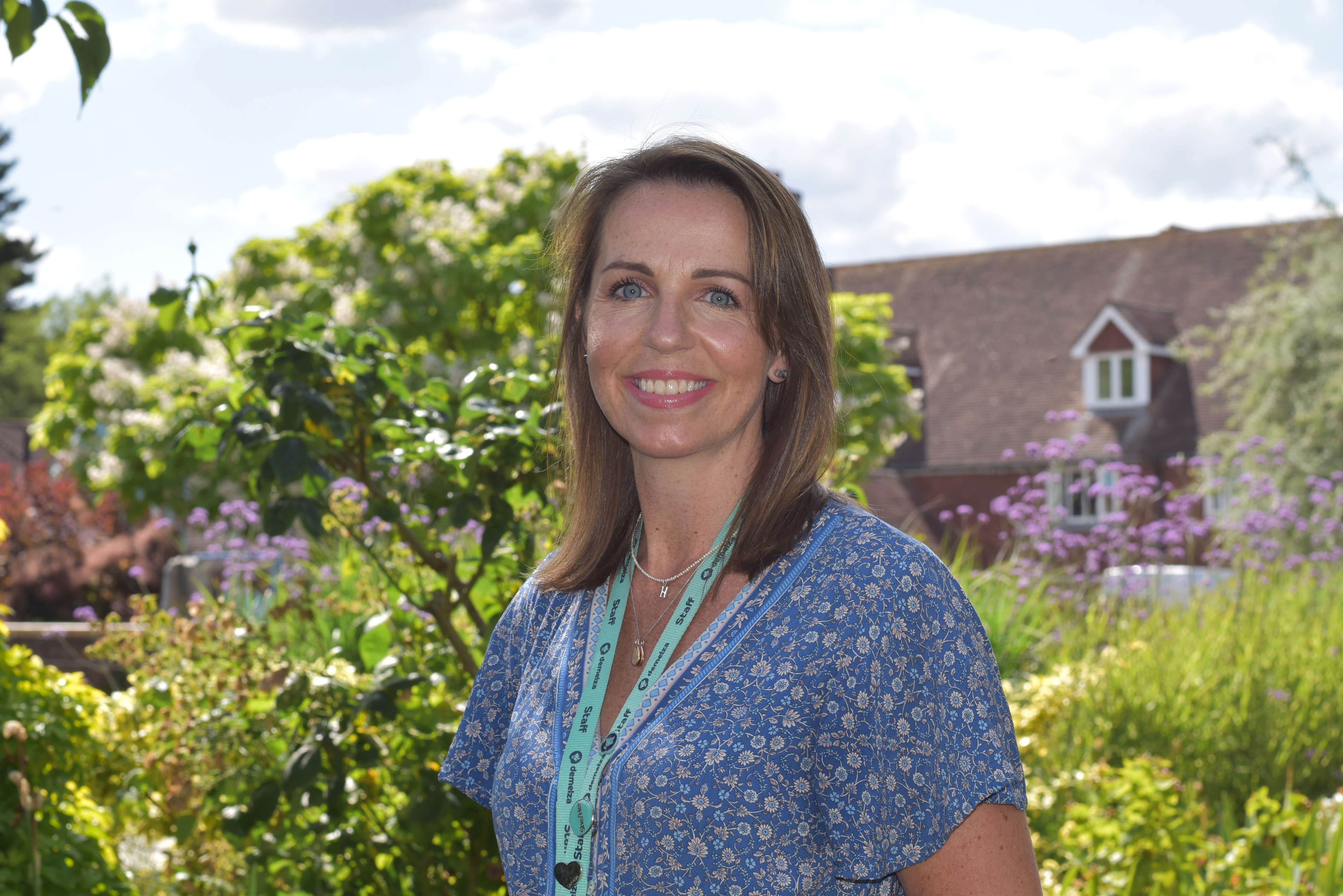 Deputy CEO Hayley Richardson stands smiling in the sunny gardens at Demelza, wearing a bright blue outfit.