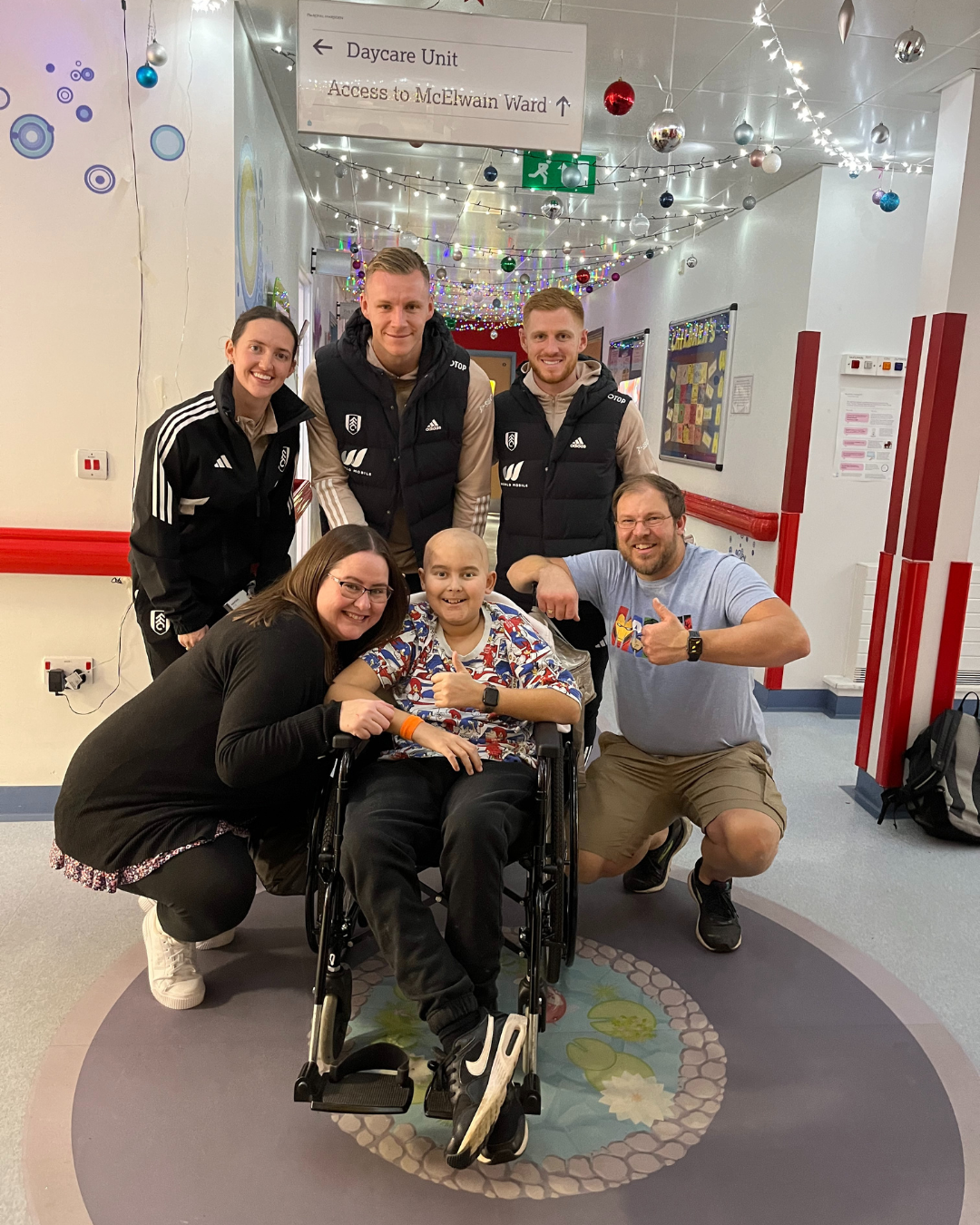 A child in a wheelchair surrounded by his parents and a few players from Fulham football club smiling towards the camera with a thumbsup