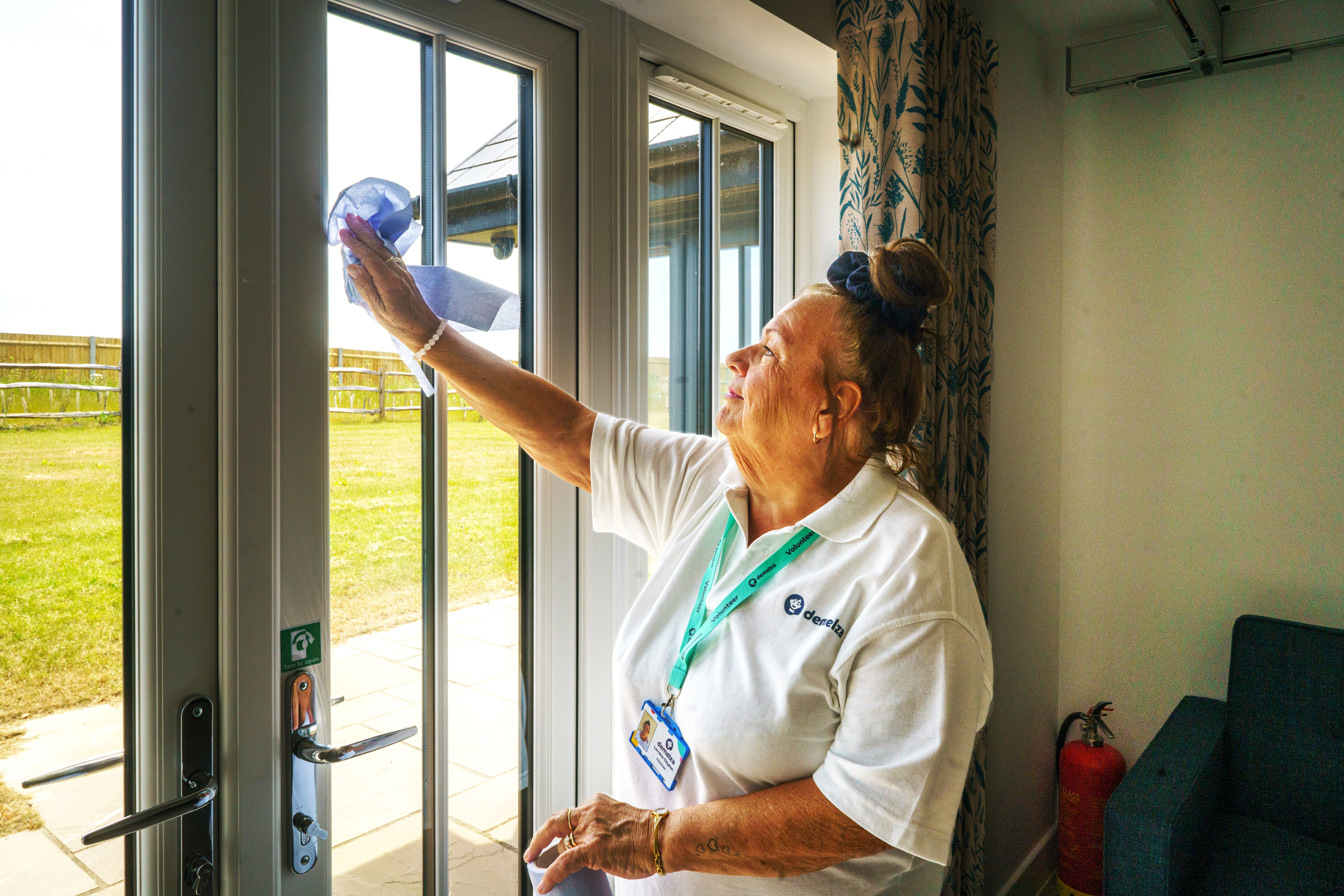 Volunteer in white uniform and lanyard, hair tied back, cleaning glass doors overlooking an outdoor area.