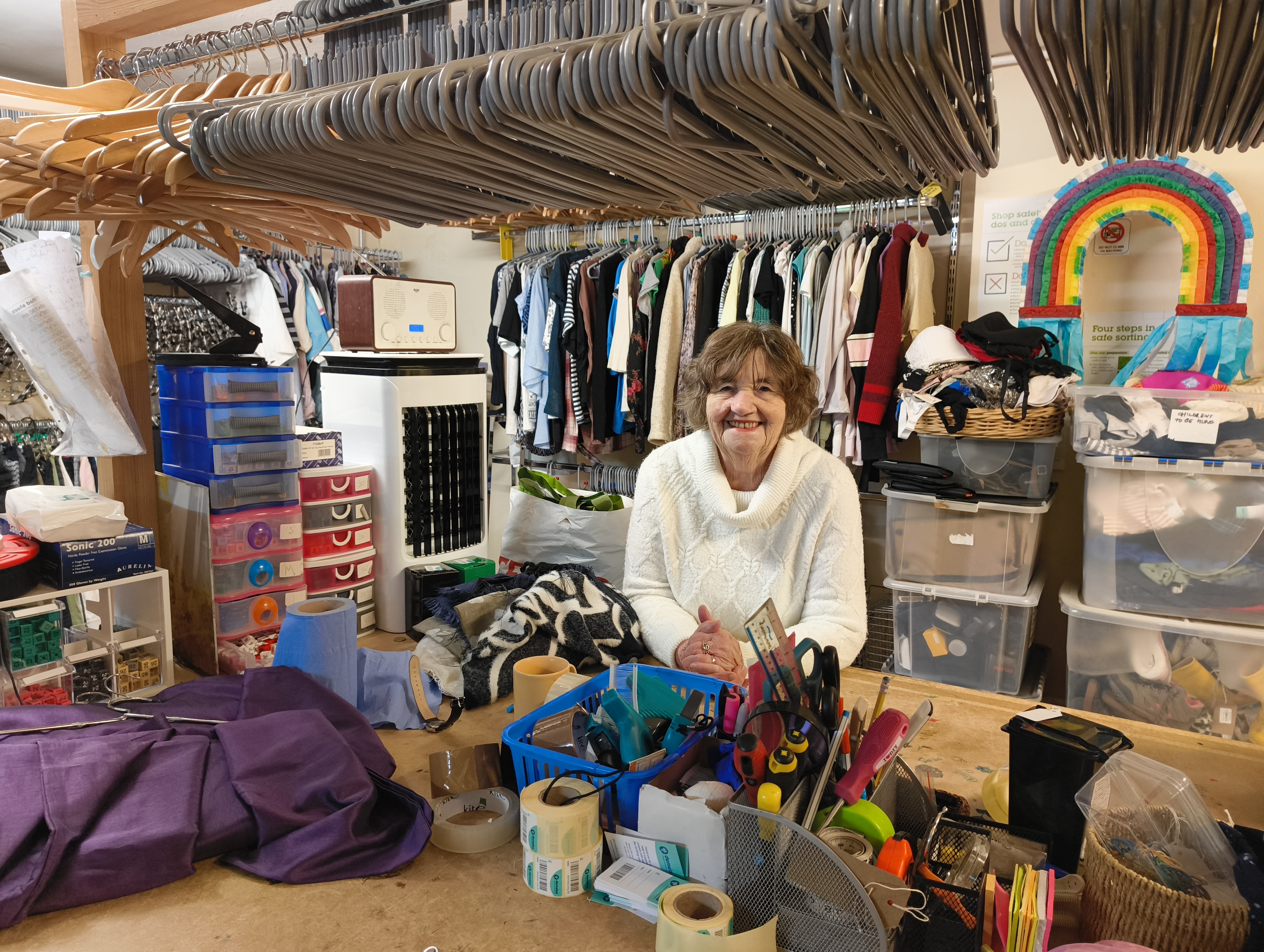 Woman stands behind a cluttered workbench in a Demelza shop with clothing racks, hangers, storage bins, and scattered tools and materials.