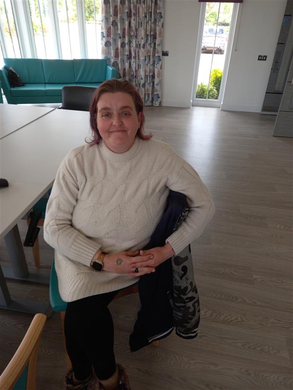 Katrina, a bereaved grandmother, sits in a chair with a gentle half-smile, reflecting quietly.