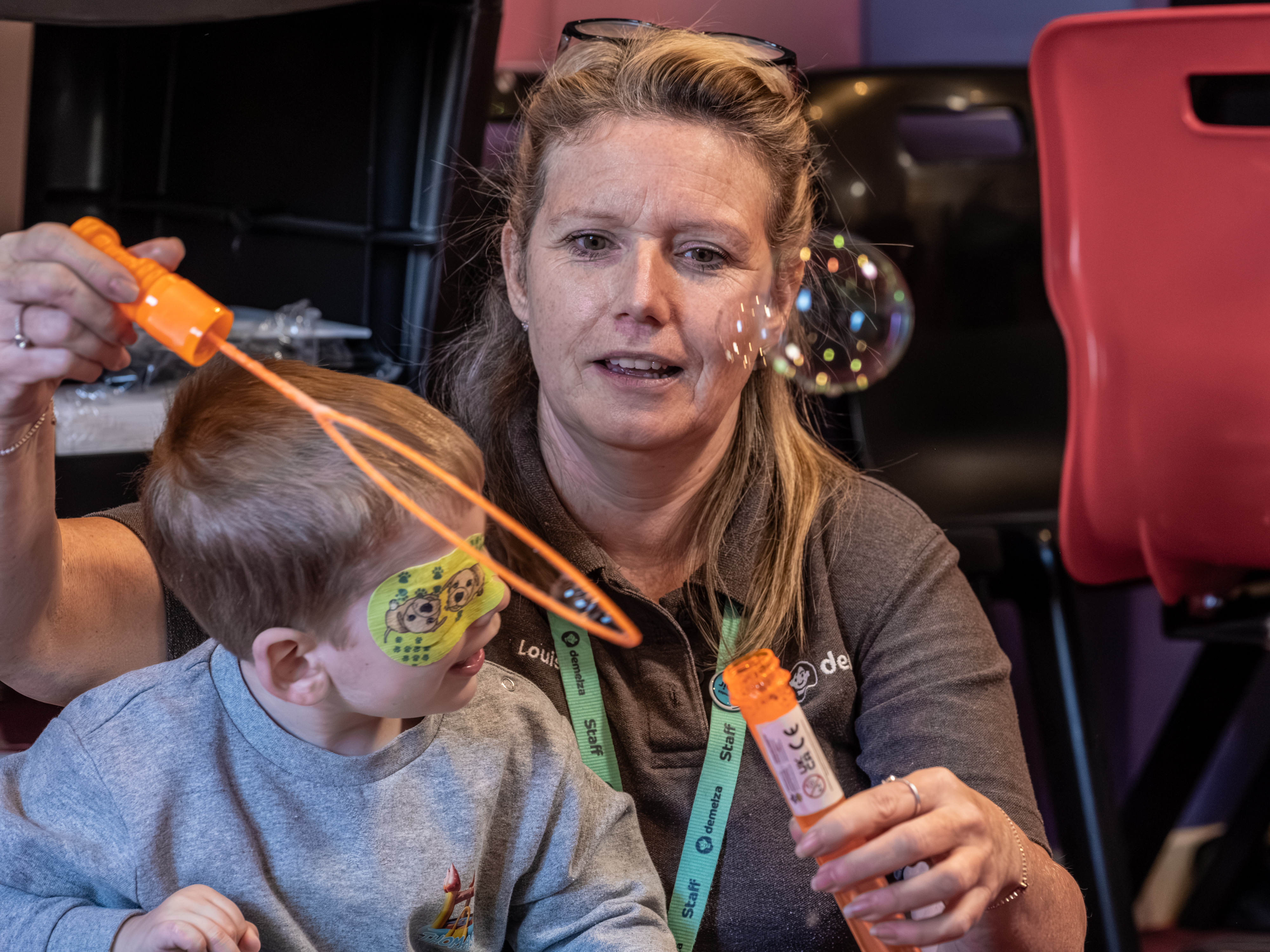 A woman with glasses on her head blowing bubbles with a young child who has a plaster over his eye