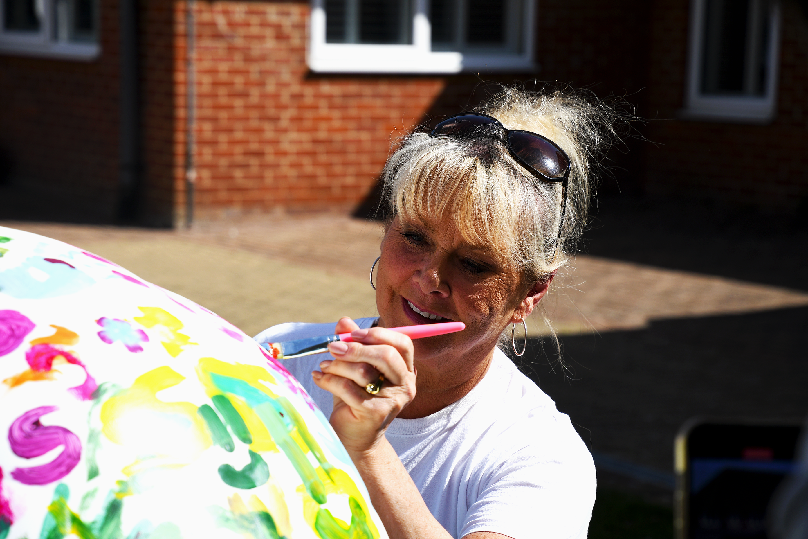 Woman with sunglasses on head painting a colourful Demelza sculpture outdoors, with a brick building visible in the background.