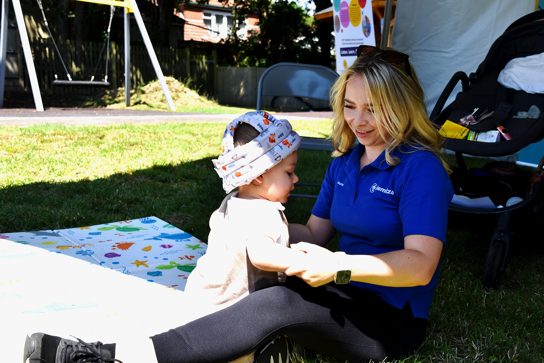 A nurse with blonde hair and in a blue Demelza shirt playing with a baby in the garden