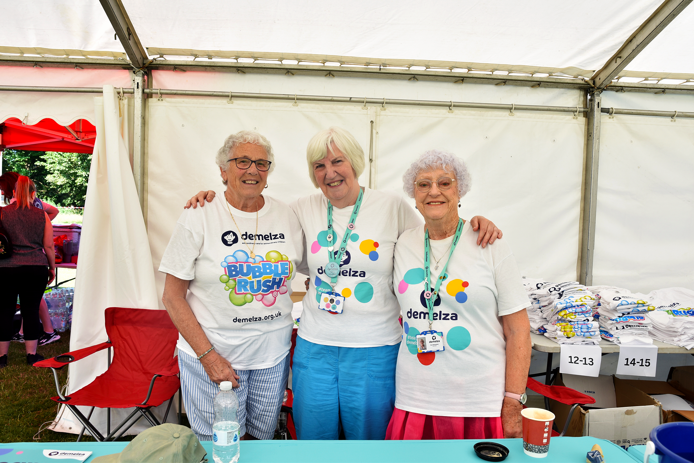 Three volunteers in bright clothes stand behind a table in a white marquee at the Mote Park Bubble Rush 2025 event.