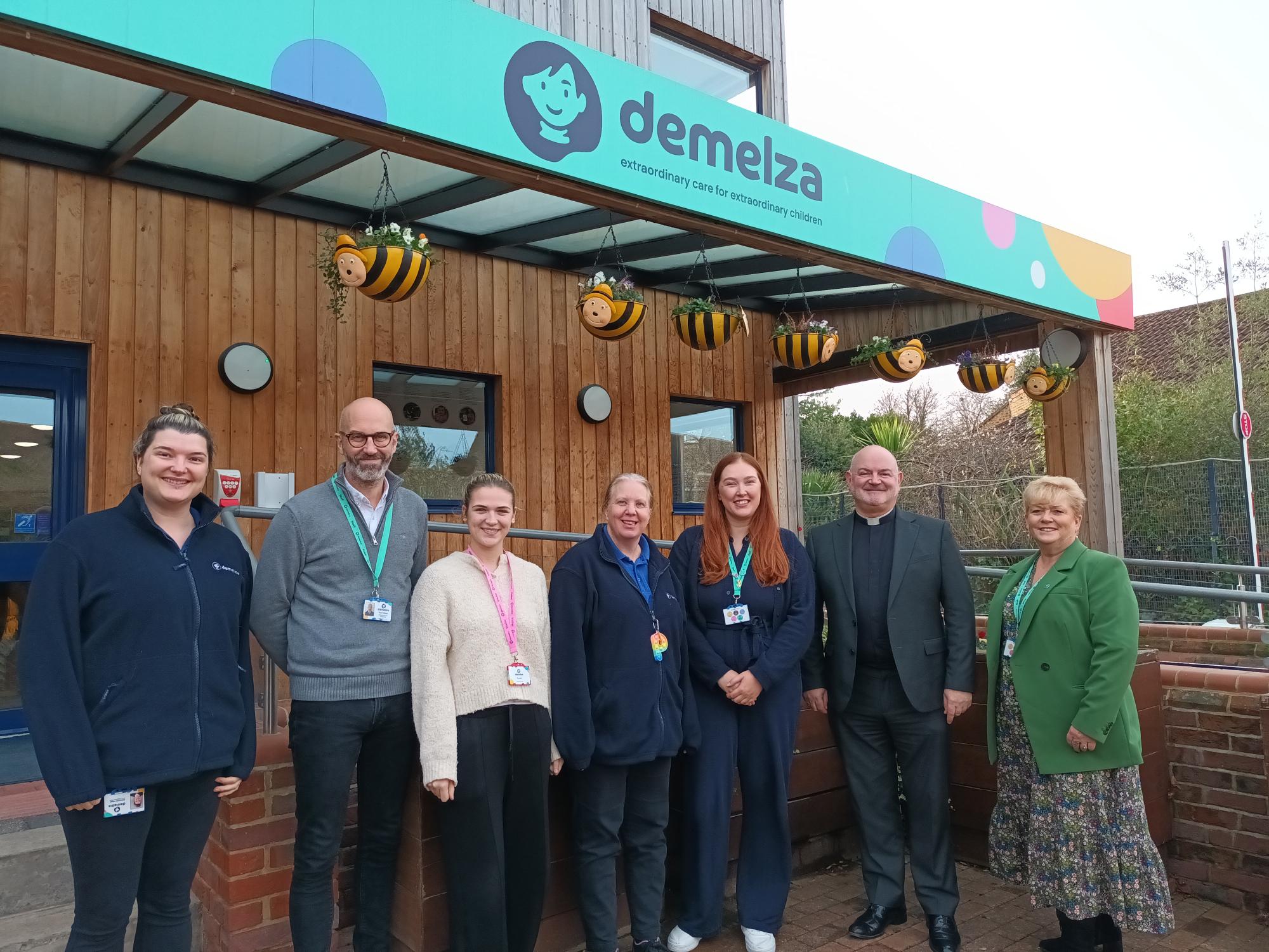Seven people stand in front of a building with a 'demelza extraordinary care for extraordinary children' sign and bee decorations hanging from the roof.