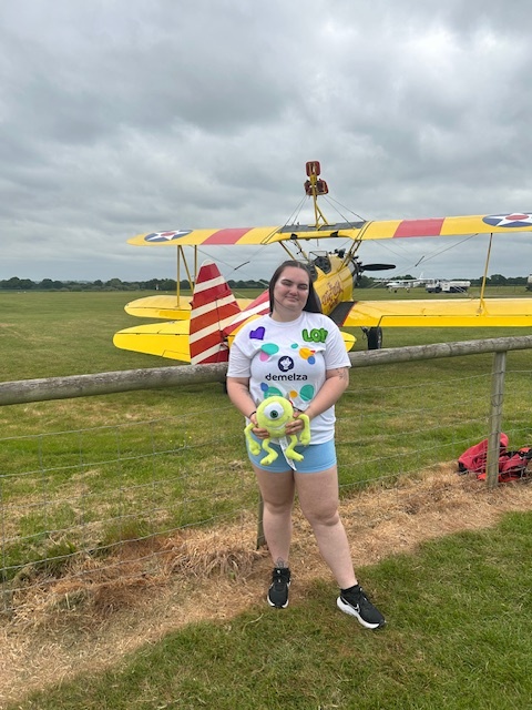 A woman holding a plush toy standing infront of a yellow plane smiling towards the camera