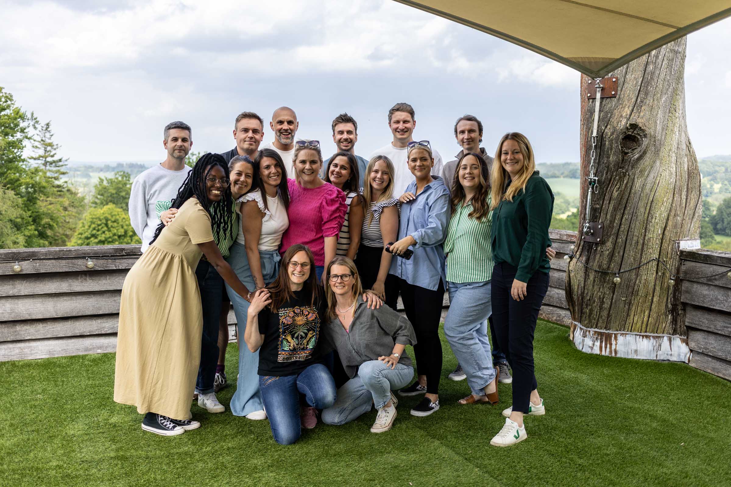 Group of fifteen people pose on grass with scenic view, wooden structure and railing behind them, under a partly cloudy sky.
