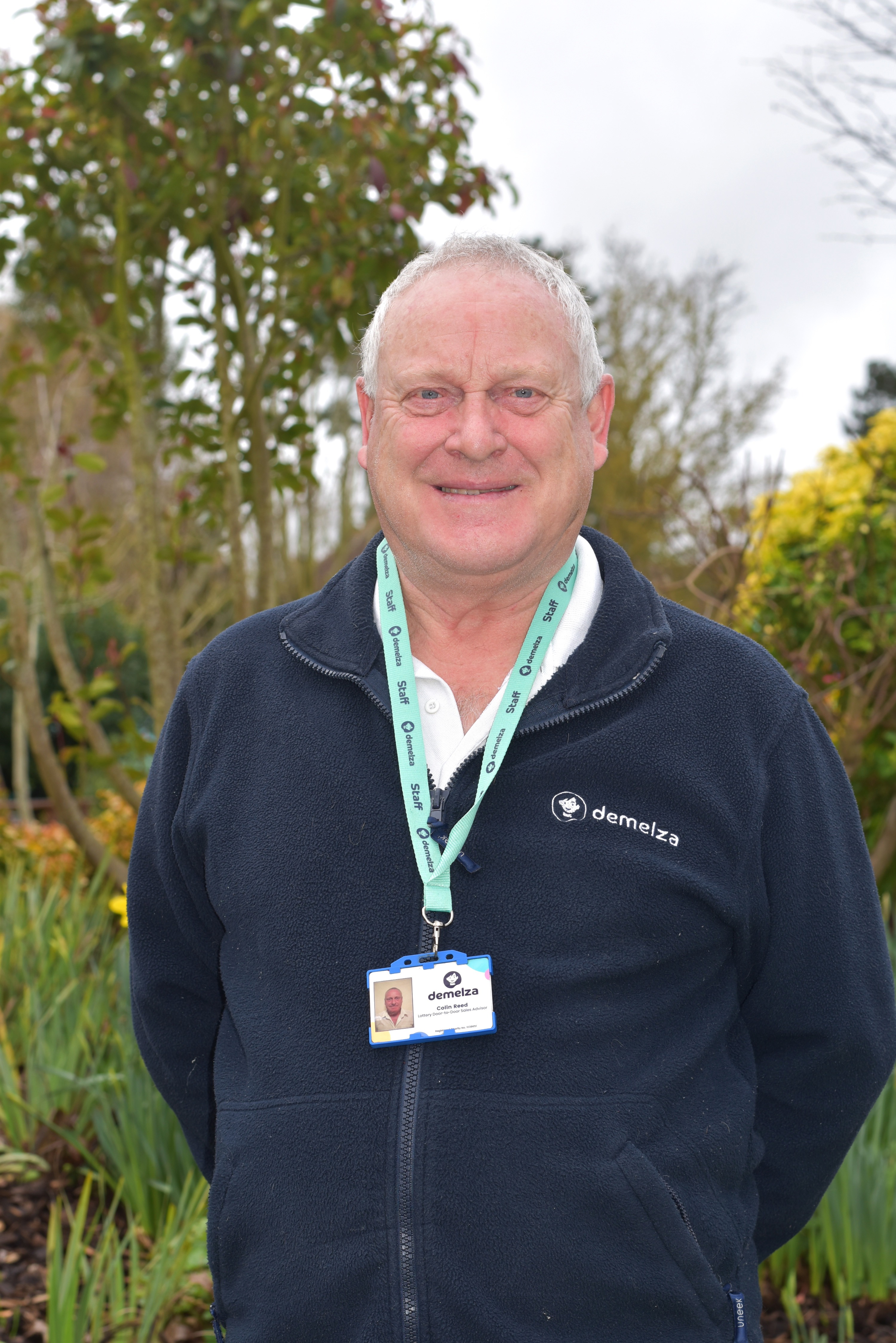 A man with white hair wearing a blue Demelza jacket outside in the tranquility garden smiling at the camera