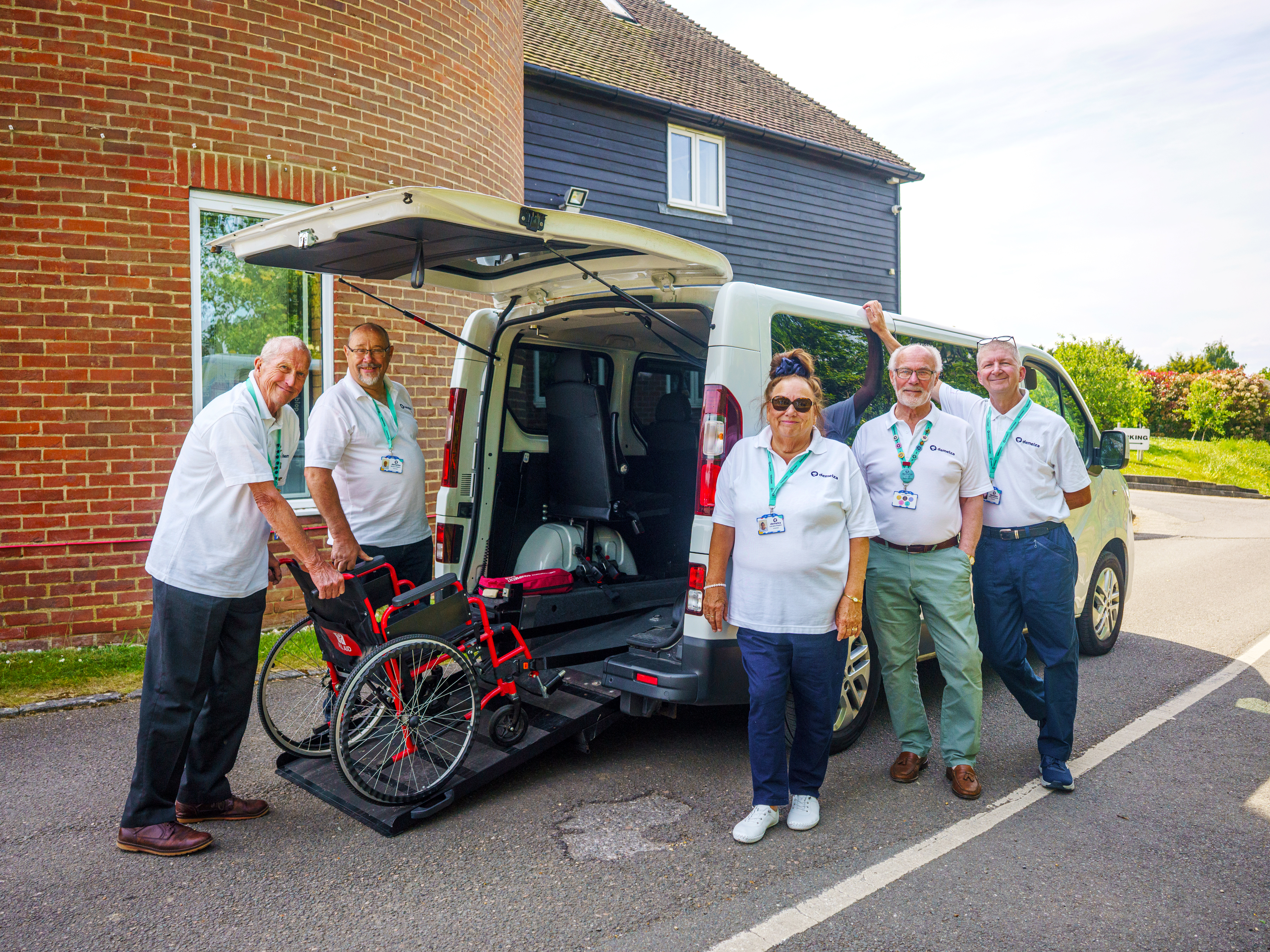 Five volunteer drivers in white uniforms stand by a minibus with its tail lift extended for wheelchair access.