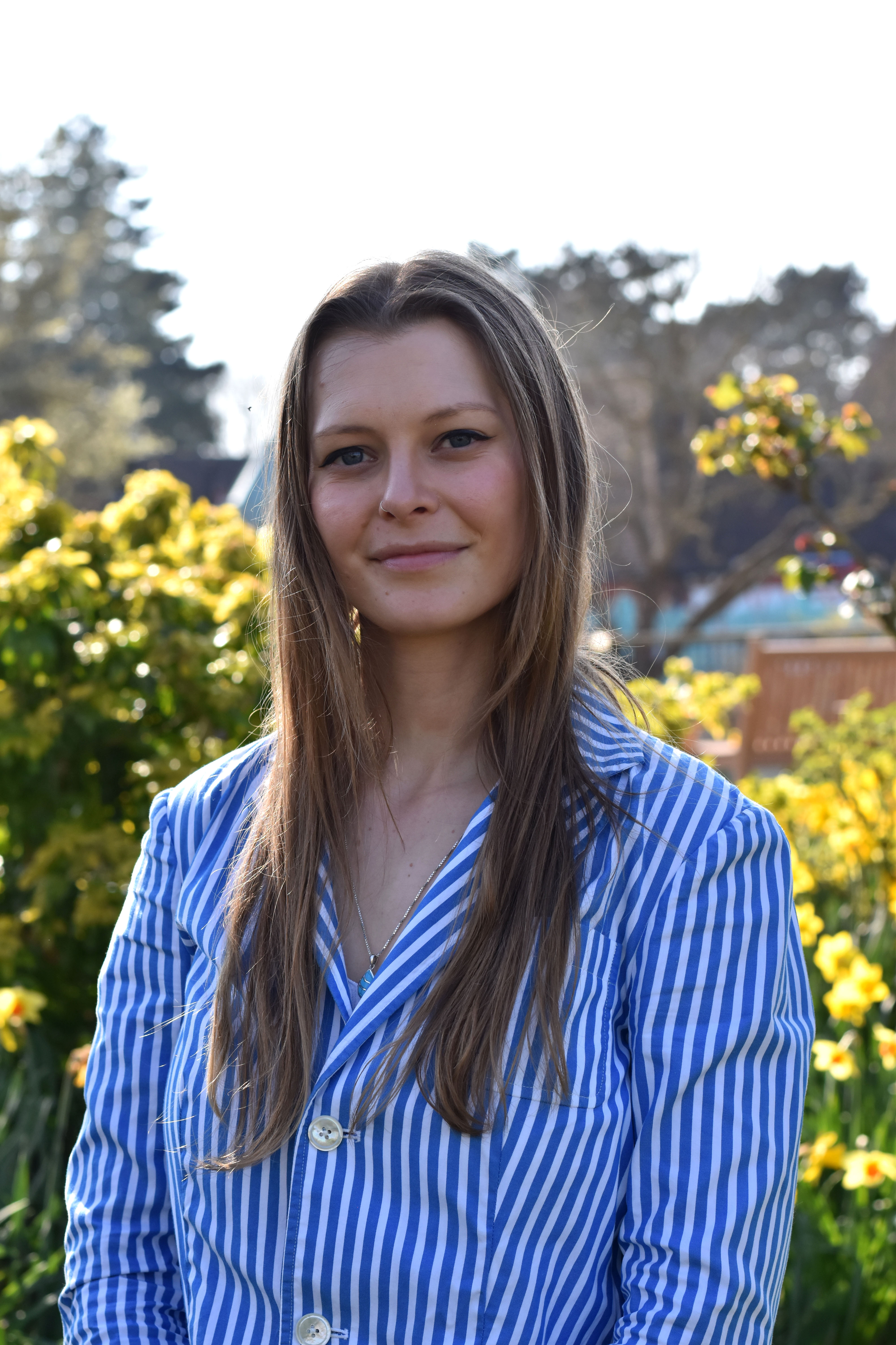 Associate Trustee Lucille Harvey stands smiling in sunny gardens, wearing a blue stripy shirt.