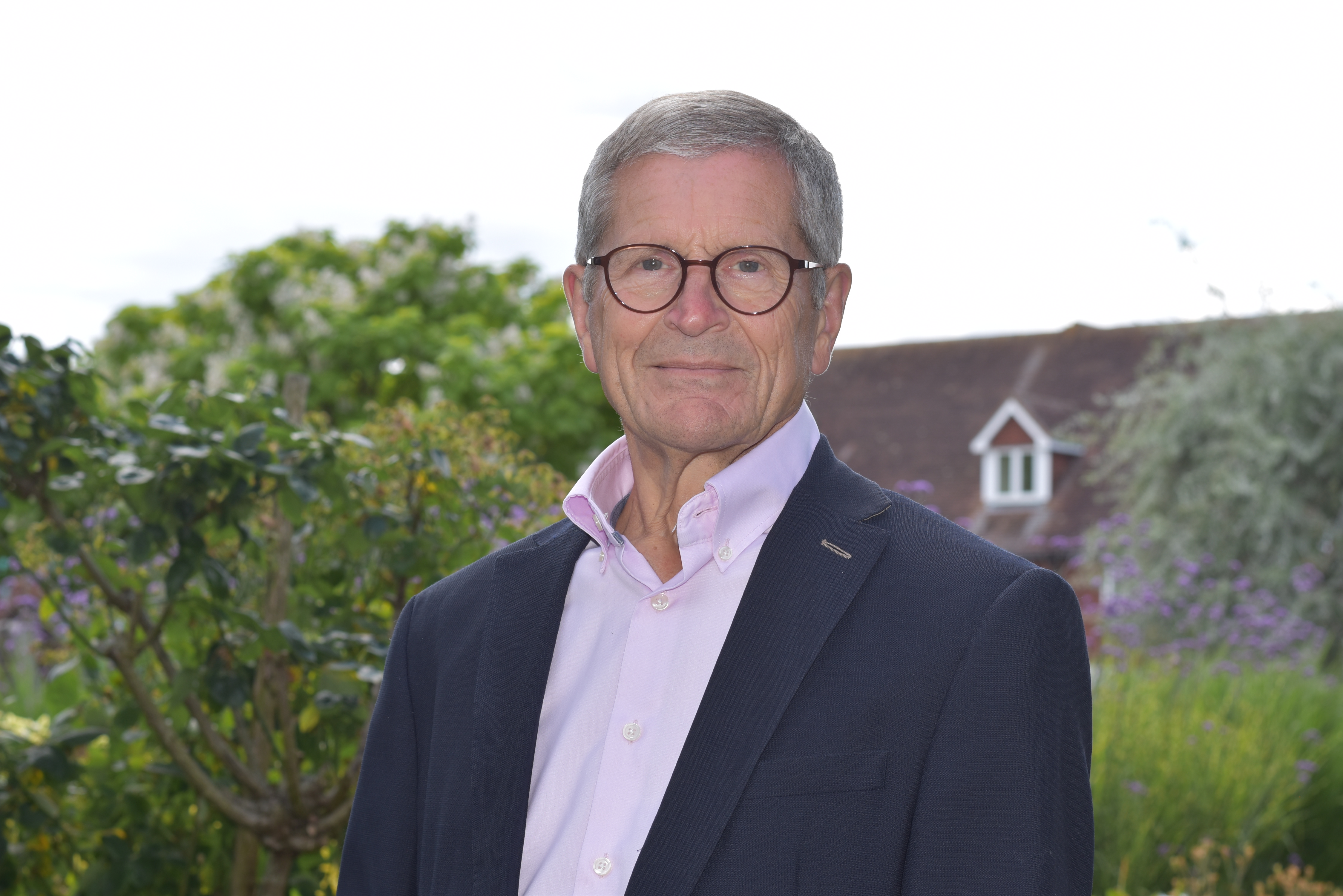 Trustee Richard Finn stands proudly in sunny gardens, wearing glasses, a pink shirt and navy blue blazer.