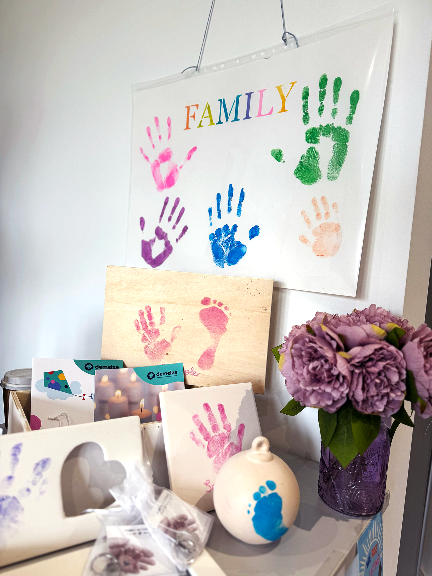 Colourful handprints and footprints labelled 'FAMILY' on a wall, with prints on wooden boards and cards. A vase of purple flowers sits nearby.