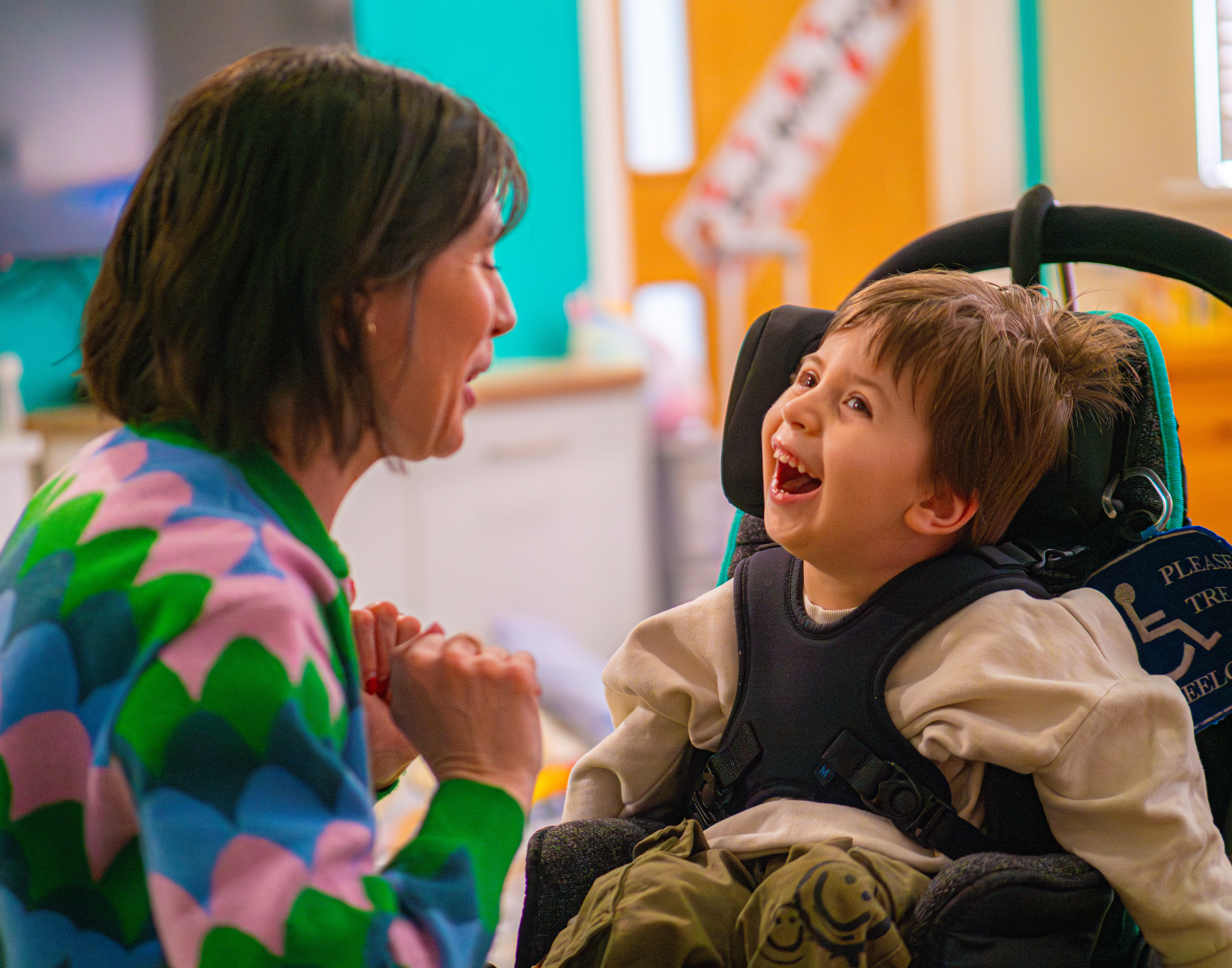 A boy in a light brown shirt in a wheelchair looking toward his mum smiling 