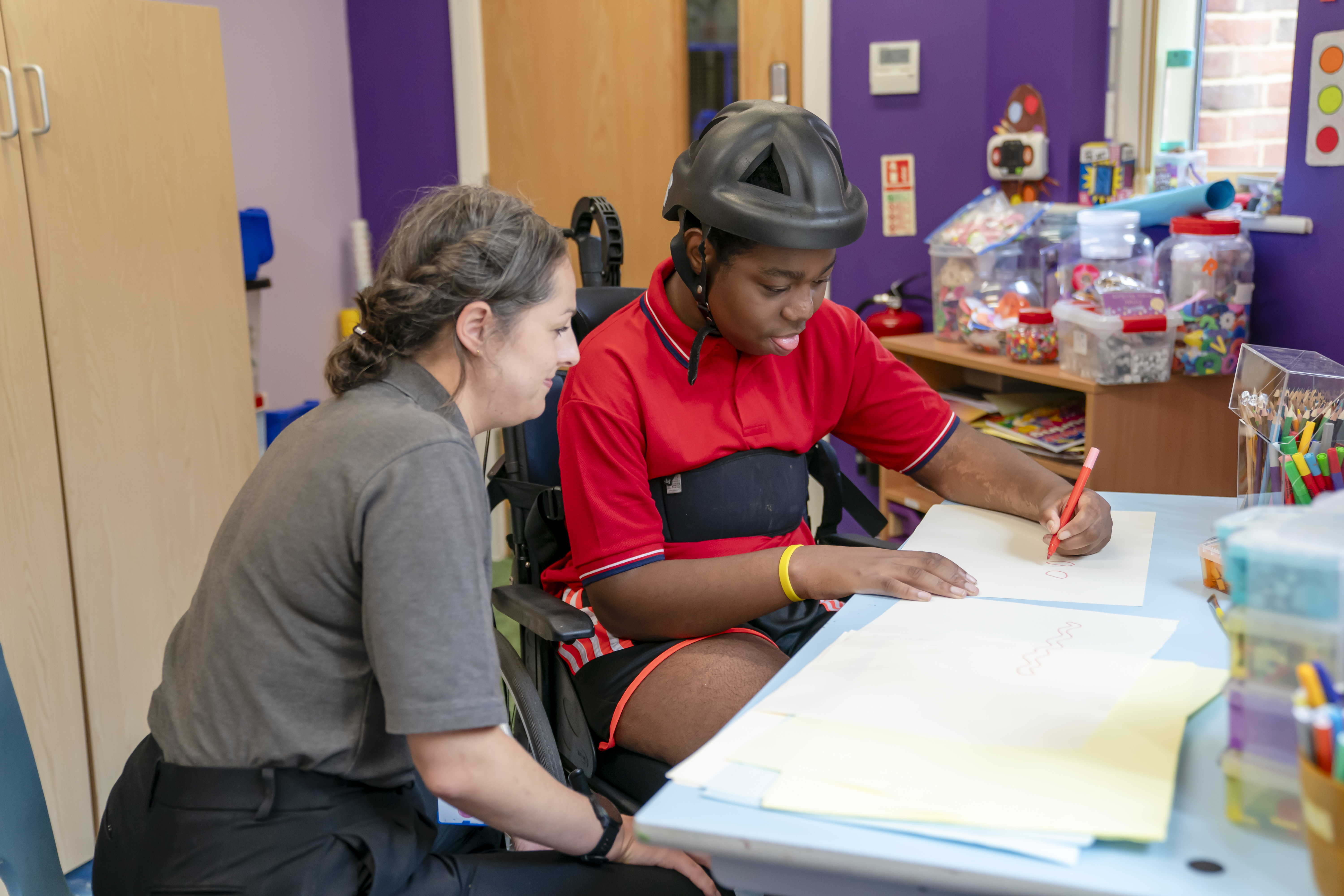 A young boy with a helmet drawing alongside a Demelza nurse