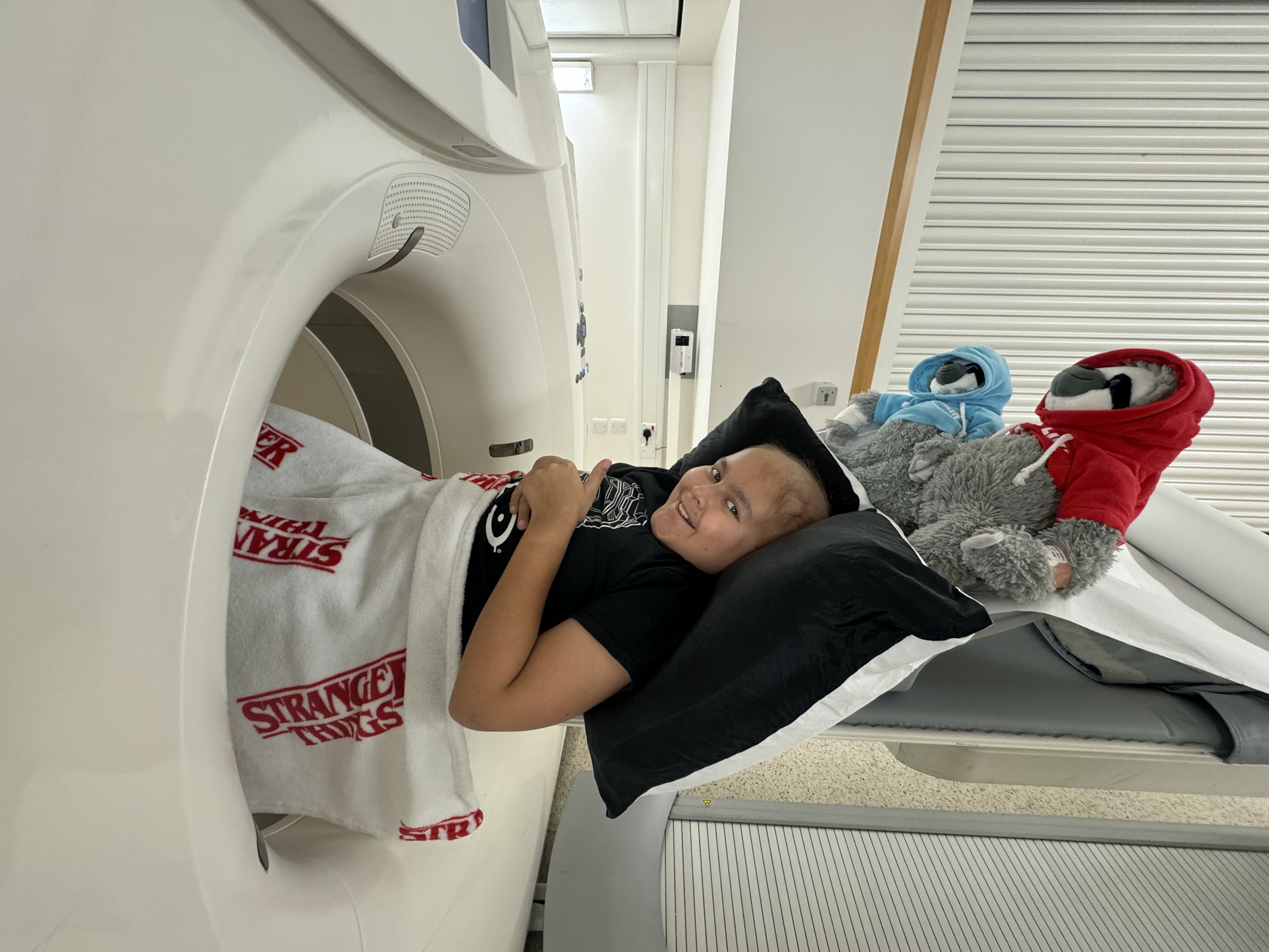 A child in a black t-shirt undergoing chemotherapy with two plush toys smiling towards the camera with a thumbs up