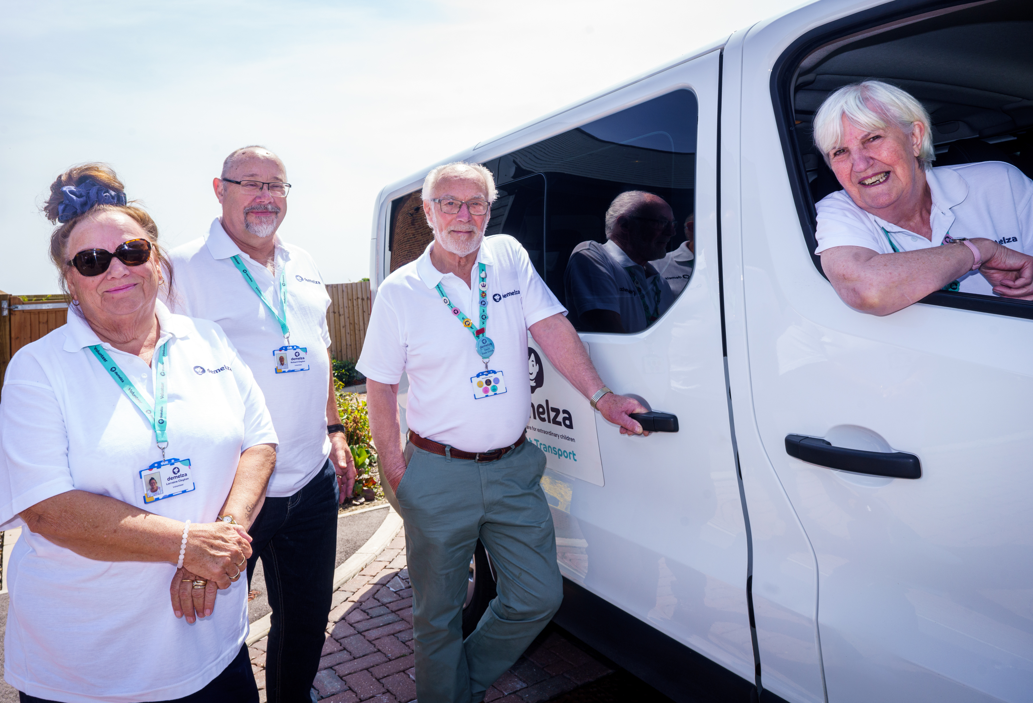 Three volunteer drivers in white uniforms stand by a white minibus, with one seated behind the wheel.