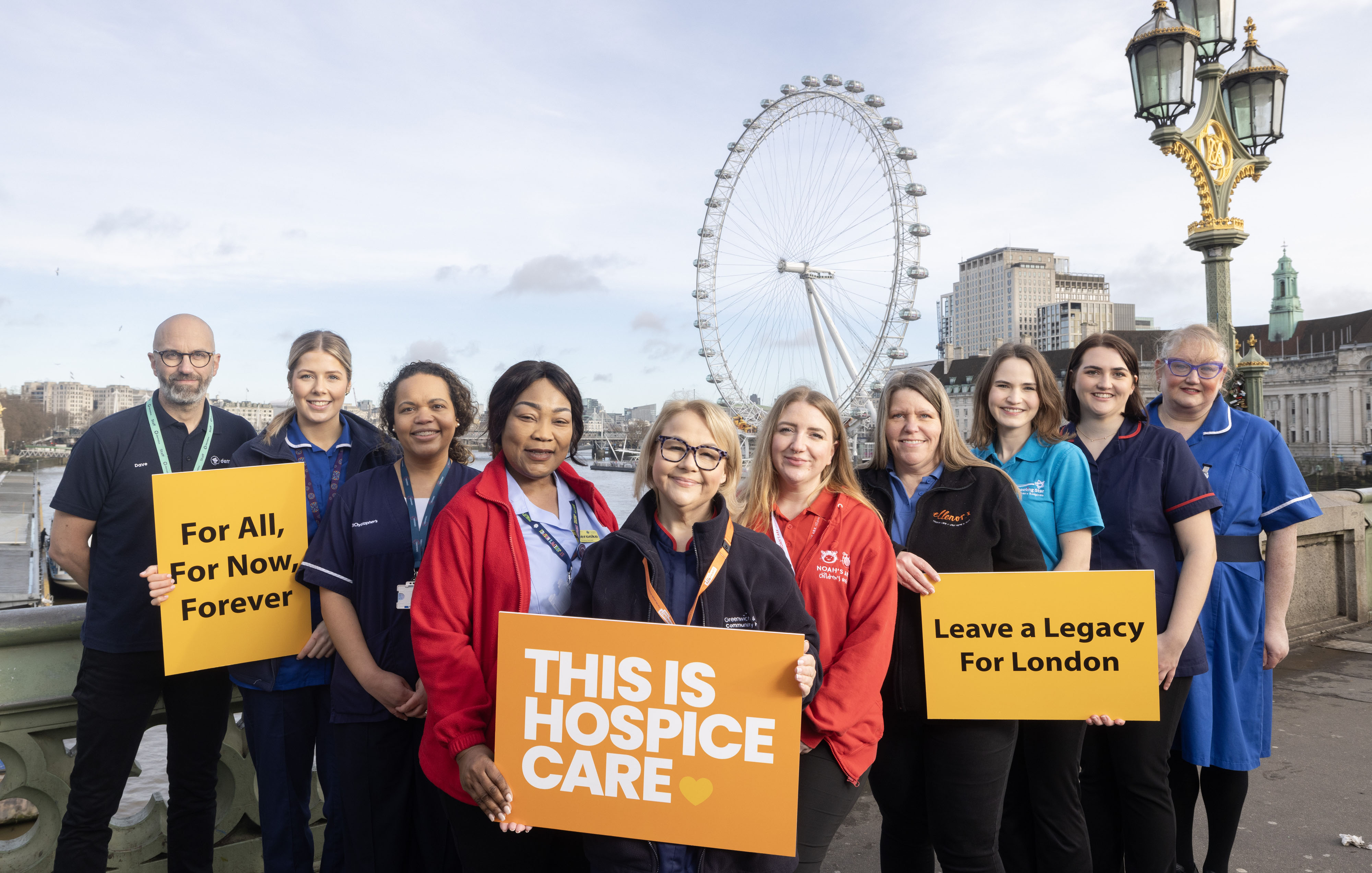 Group of people stand in front of the London Eye holding signs: 'For All, For Now, Forever,' 'THIS IS HOSPICE CARE,' and 'Leave a Legacy For London.'