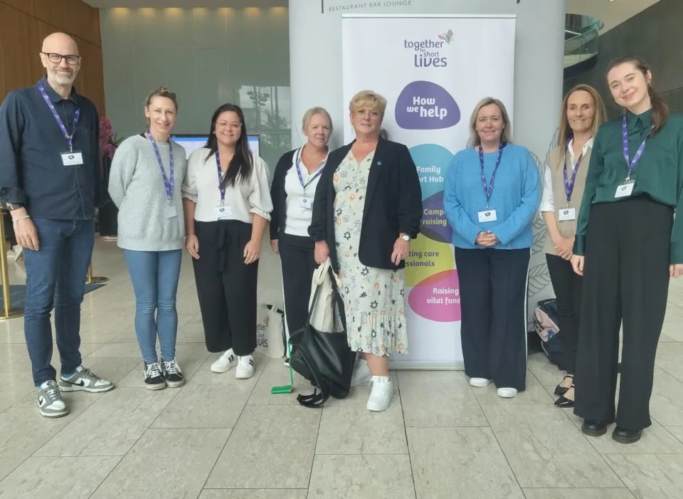 Eight people stand indoors in front of a banner reading 'together we change Lives' with sections on support, counselling, and fundraising.