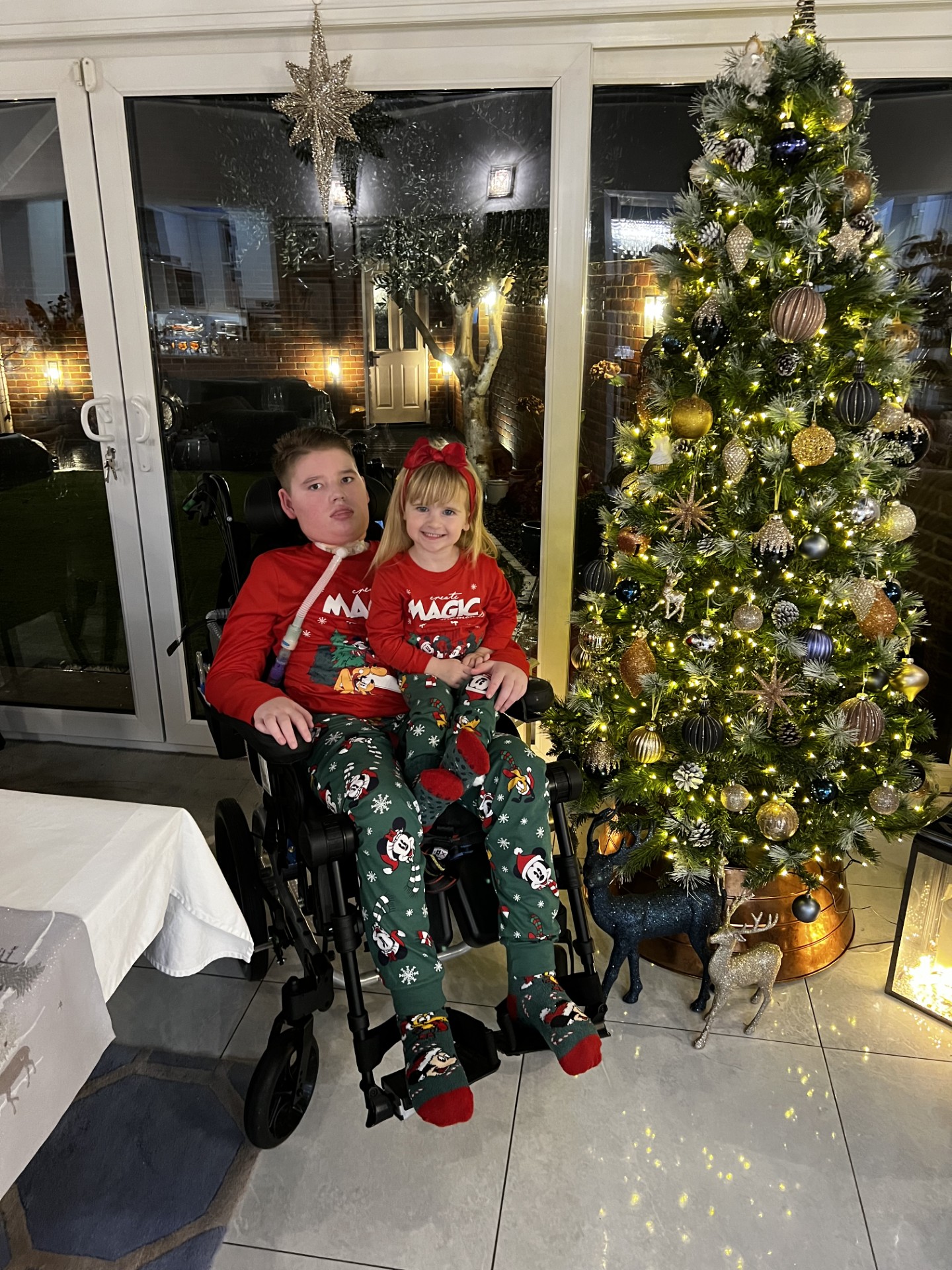 Boy on a wheelchair with his sister on his lap, both wearing christmas jumpers, , smiling whilst next to a christmas tree