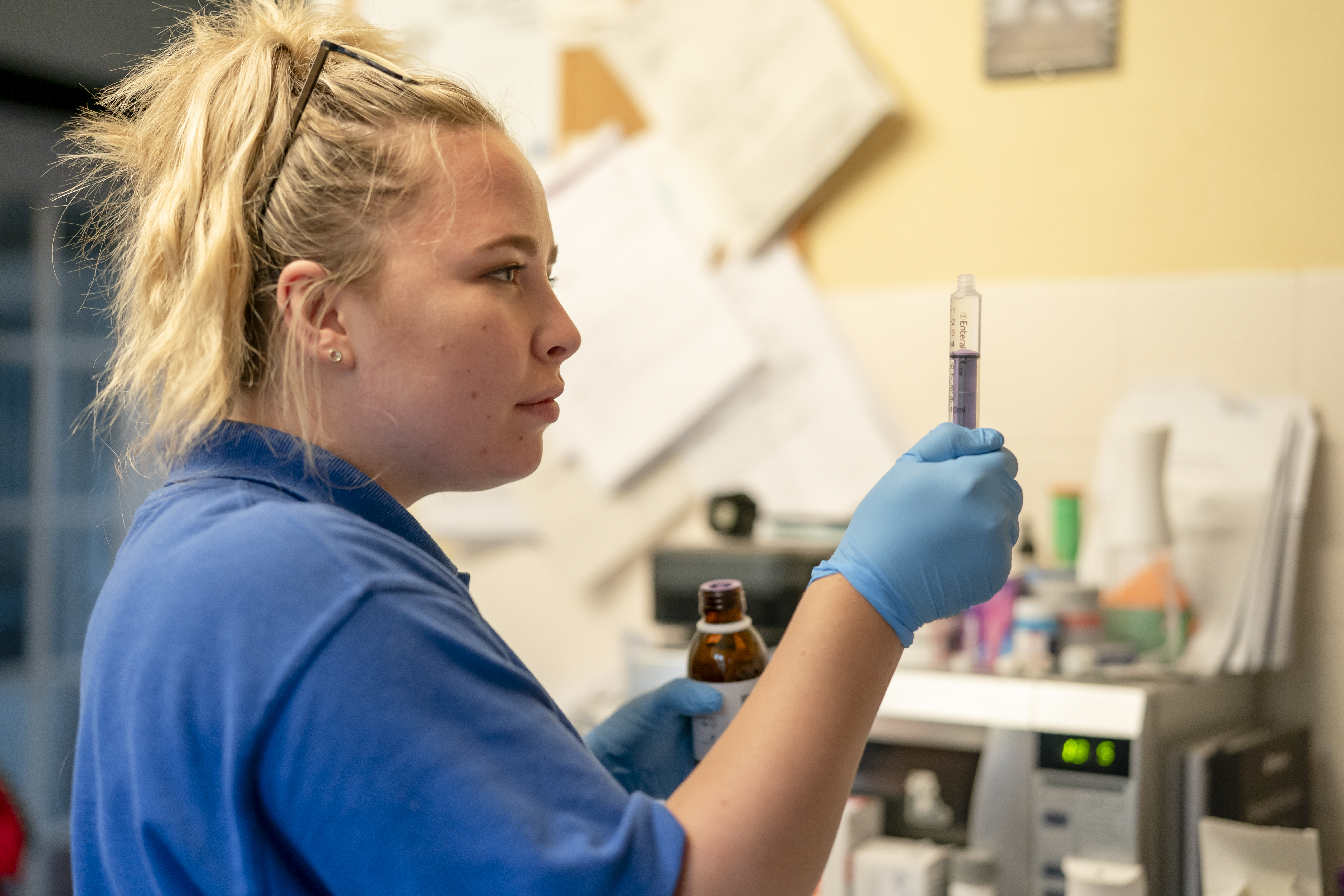 Nurse Ella, wearing a blue uniform, glasses on her head, and gloves on her hands, preparing medication.