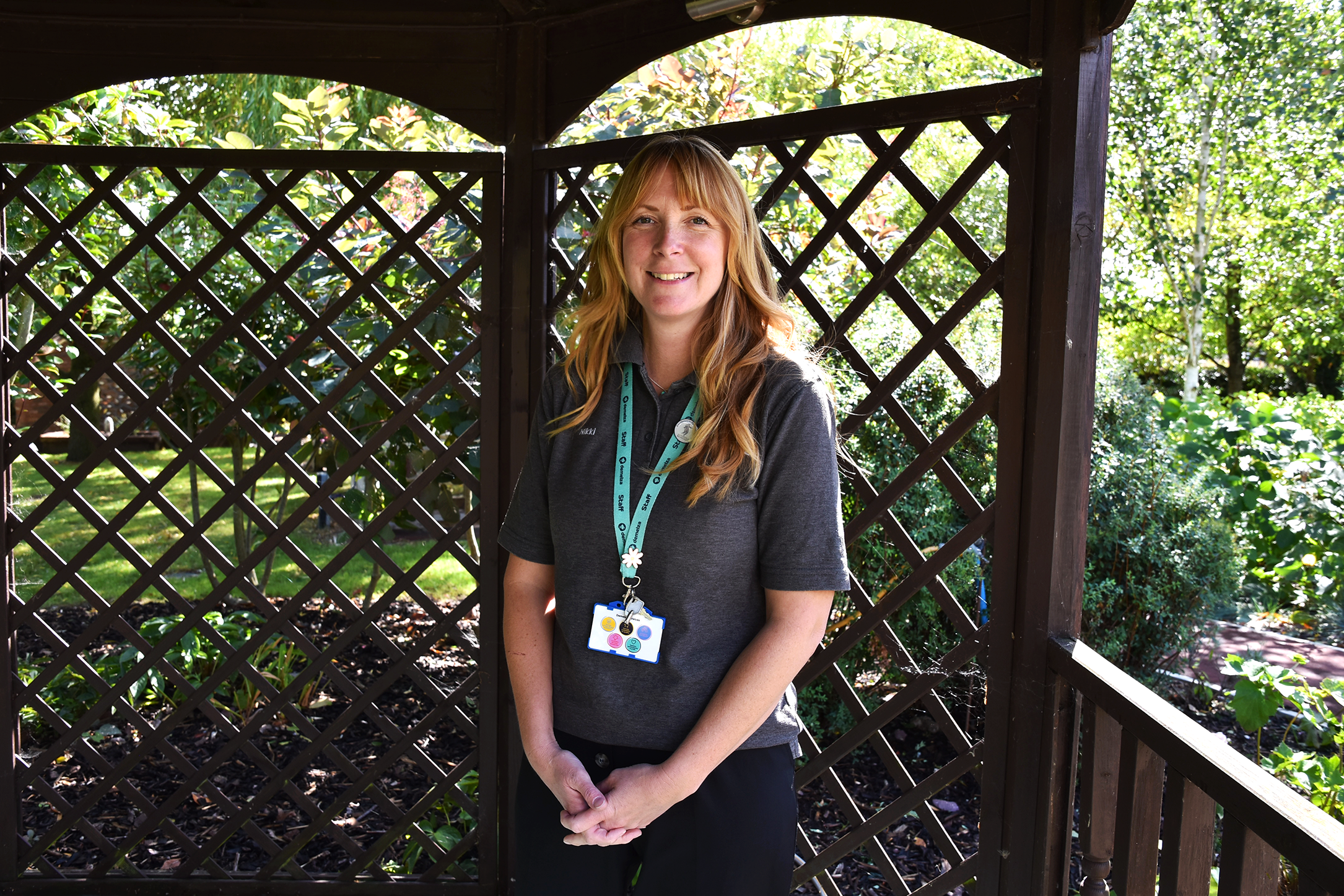 A person wearing a grey shirt and a Demelza ID badge on a lanyard stands inside a wooden gazebo with lattice walls. Greenery and trees are visible in the background.