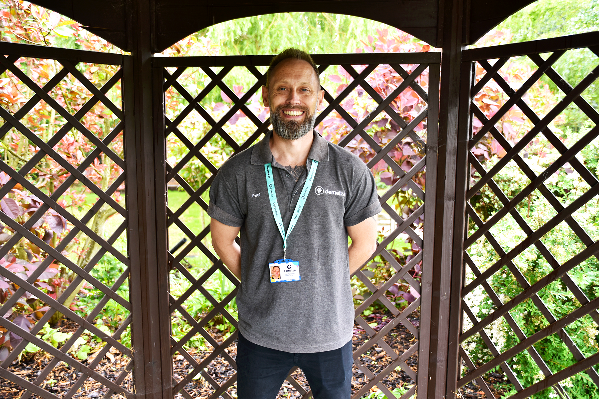 Man stands in front of wooden lattice, wearing grey polo and Demelza ID badge. Background includes greenery and plants.