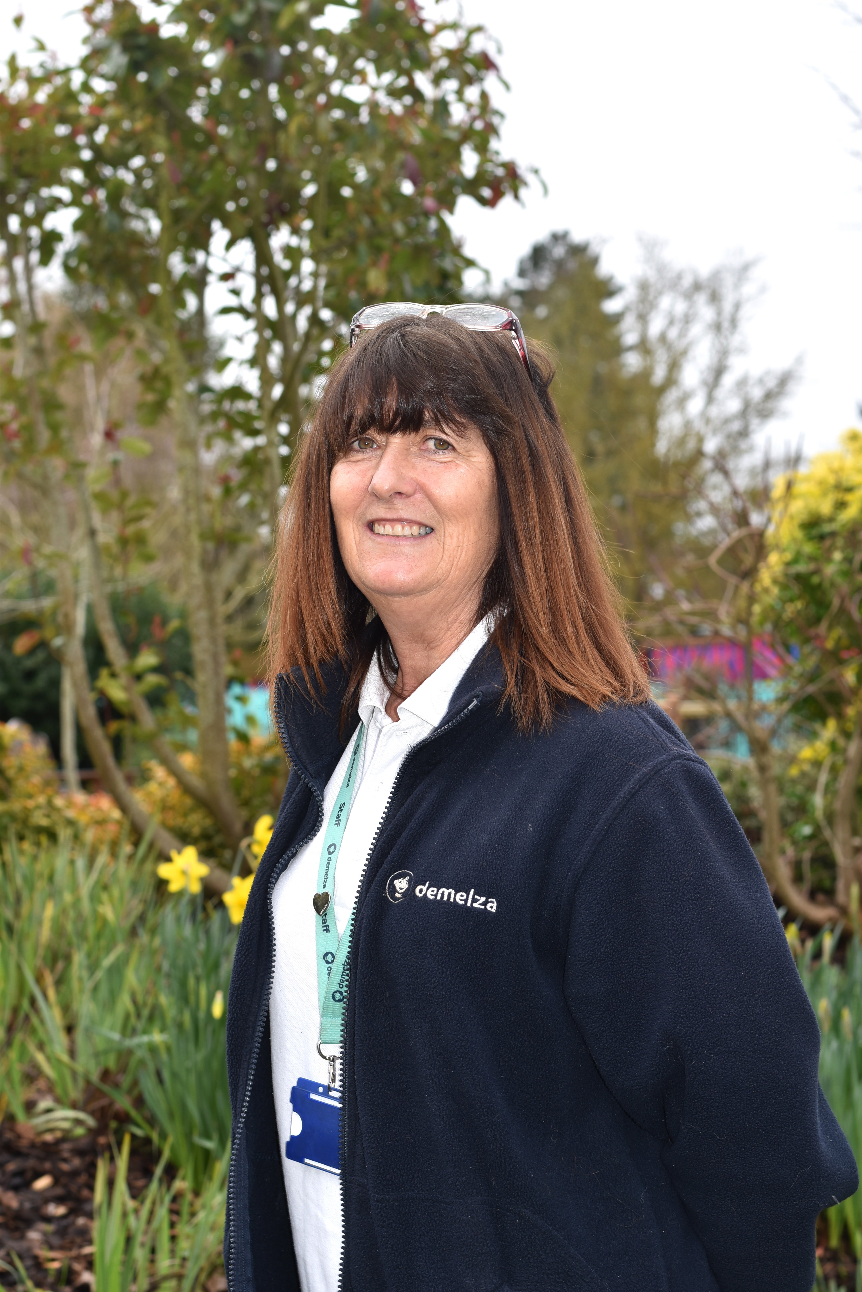 A woman with brown hair wearing a blue Demelza jacket outside in the tranquility garden smiling at the camera