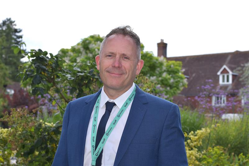 Ashley Henson, Director of Retail, stands smiling in Demelza’s hospice gardens wearing a blue suit and tie.