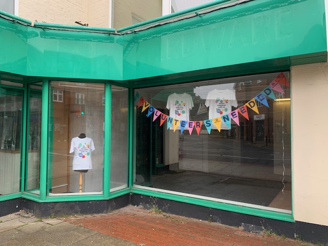 Storefront with green trim showing white Demelza t-shirts on a mannequin and inside; a colorful "VOLUNTEERS NEEDED" banner is displayed.