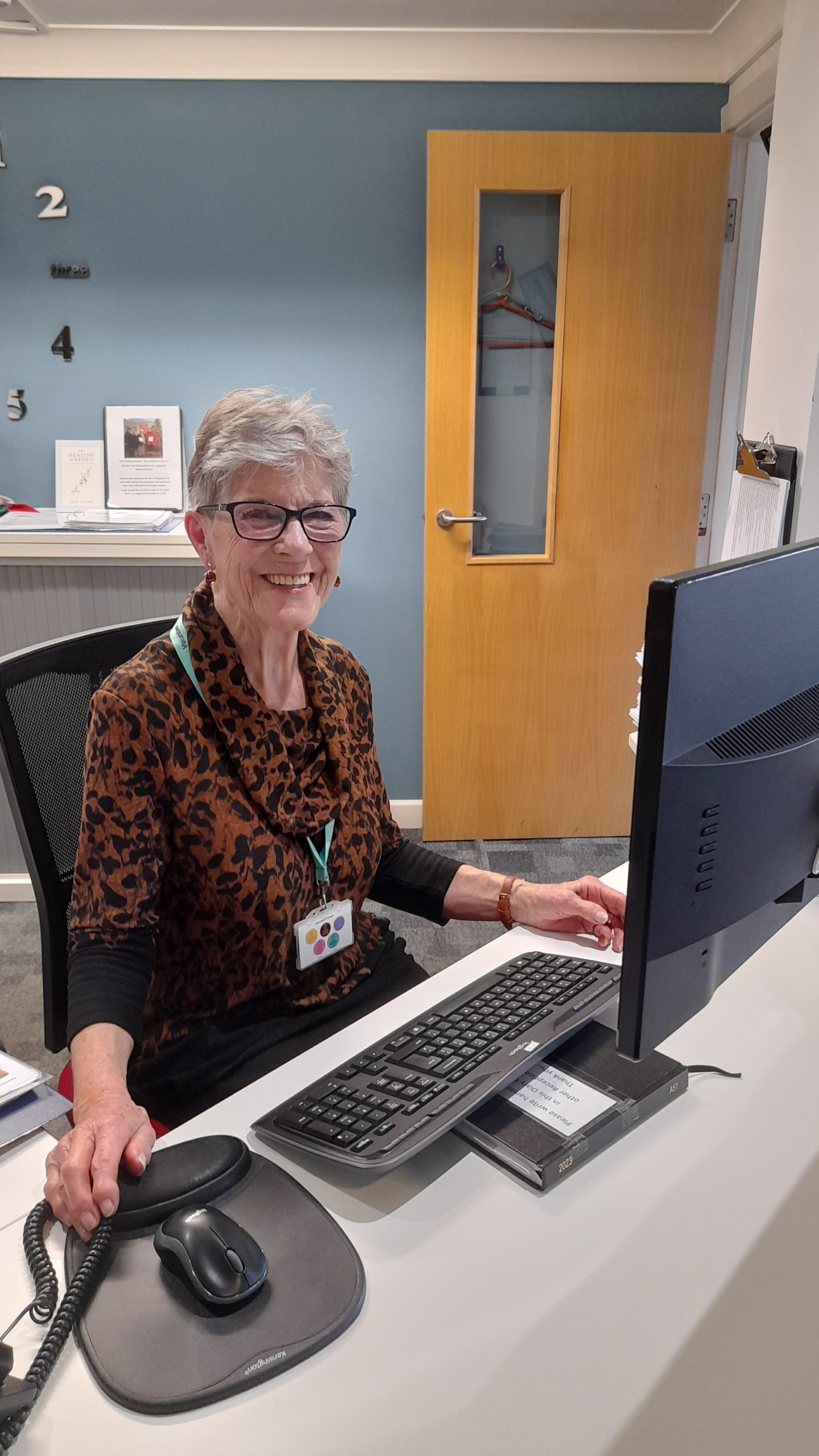 Lynn sits at a reception desk in front of a computer, smiling and ready to welcome visitors.