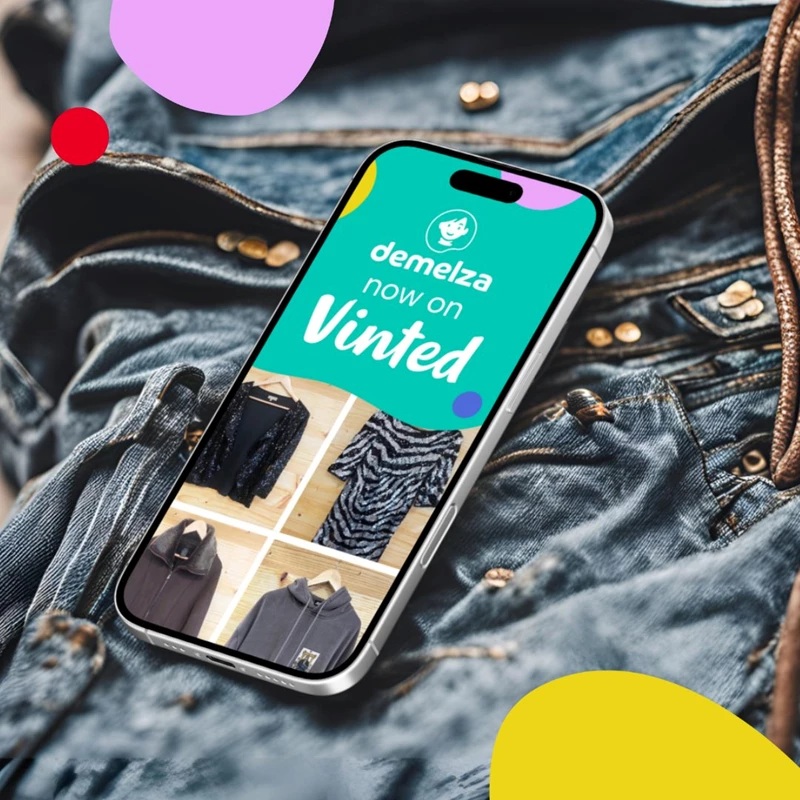 Smartphone showing 'demelza now on Vinted' with clothing images, placed on denim fabric background with colorful shapes.