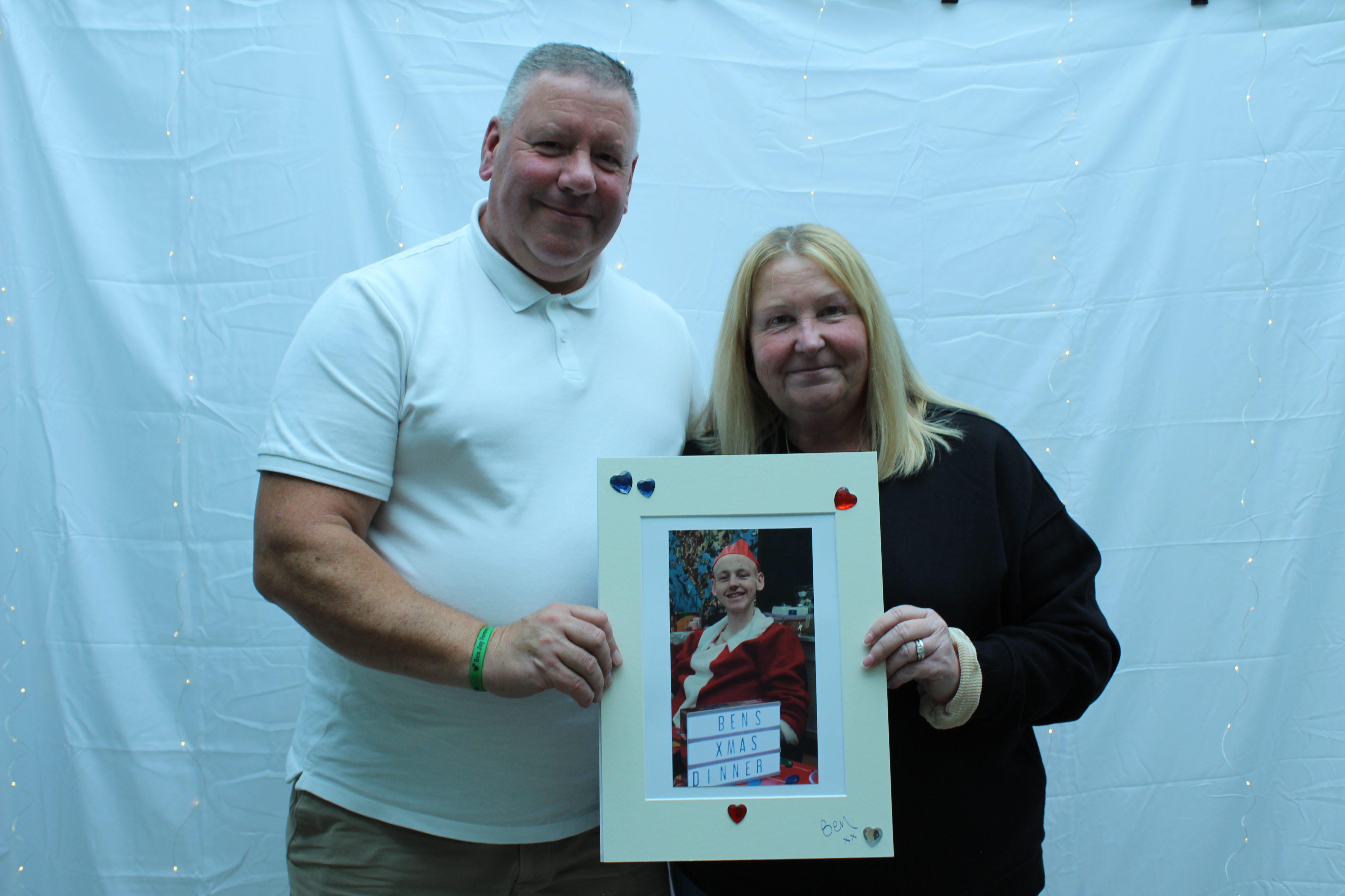 A man in a white shirt and a woman in a black shirt holding up a photo of their son who sadly passed away