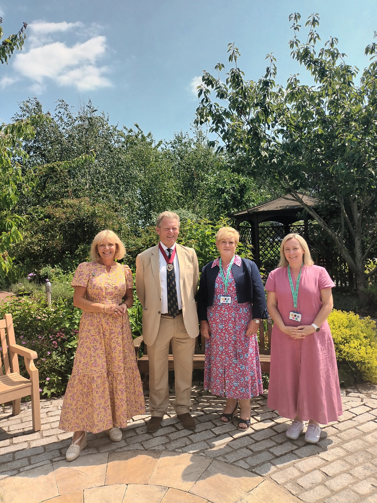 Four people with lanyards stand outdoors on a sunny day. Three wear floral dresses, one a beige suit, with greenery in the background.