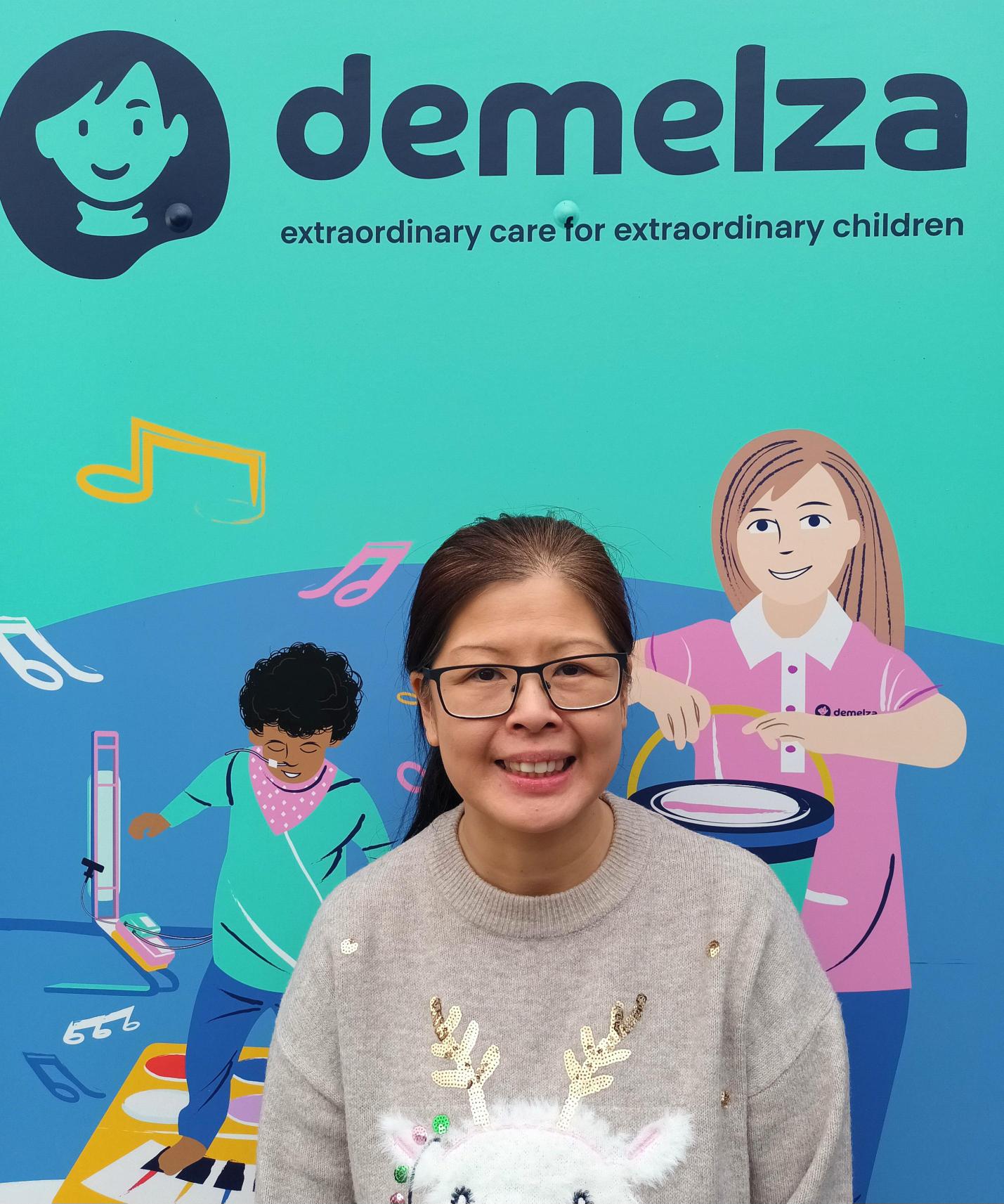 Woman stands in front of a colorful 'demelza' backdrop with the tagline 'extraordinary care for extraordinary children' and illustrations of children playing instruments.
