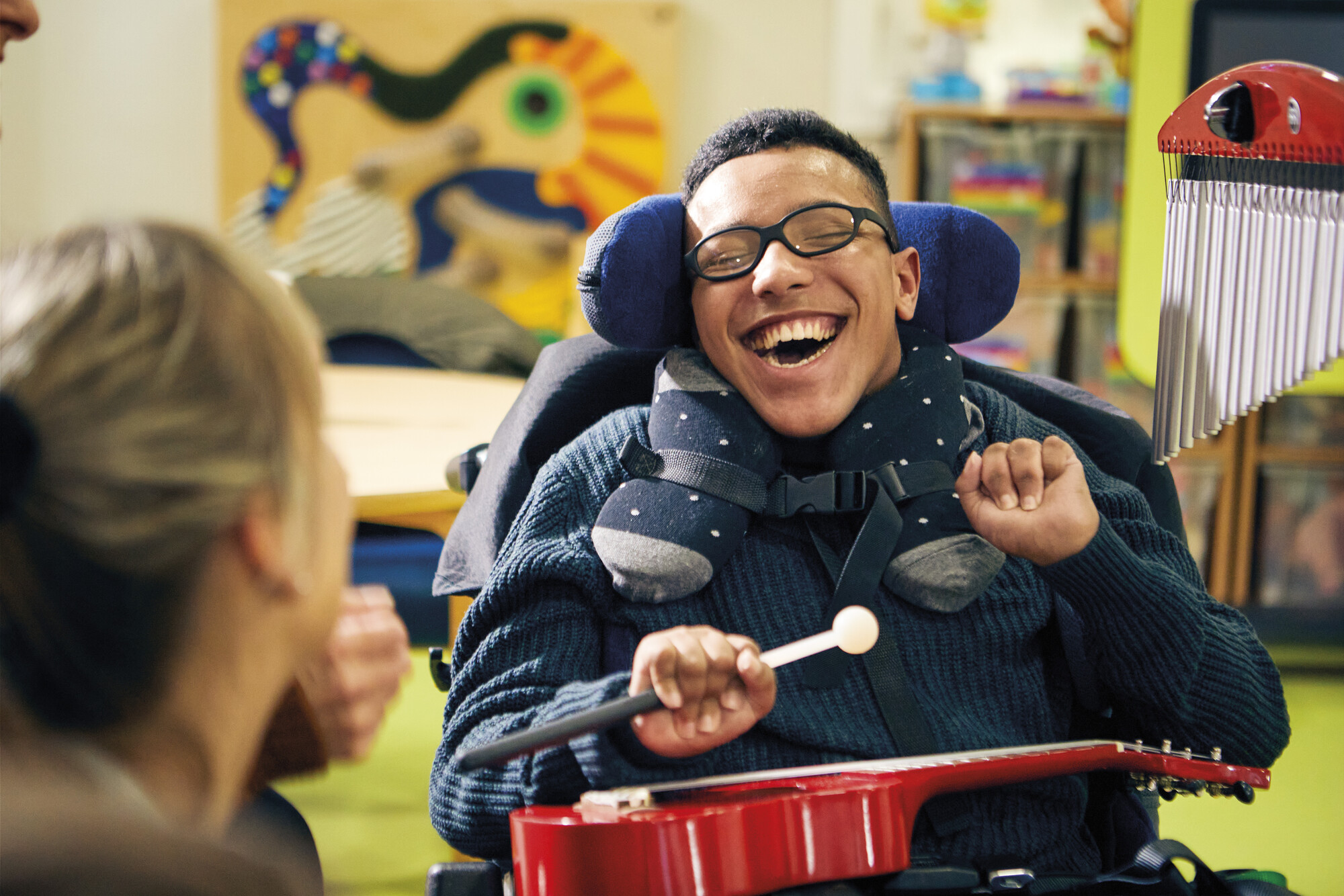 Liam enjoys music at Eltham, smiling broadly with a drumstick in hand and a guitar resting on his lap.