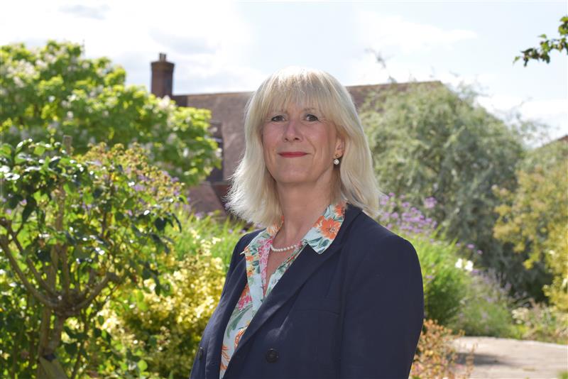 Trustee Nicola Tyers smiles proudly in sunny gardens, wearing a floral blouse, navy blue jacket and pearl jewellery.